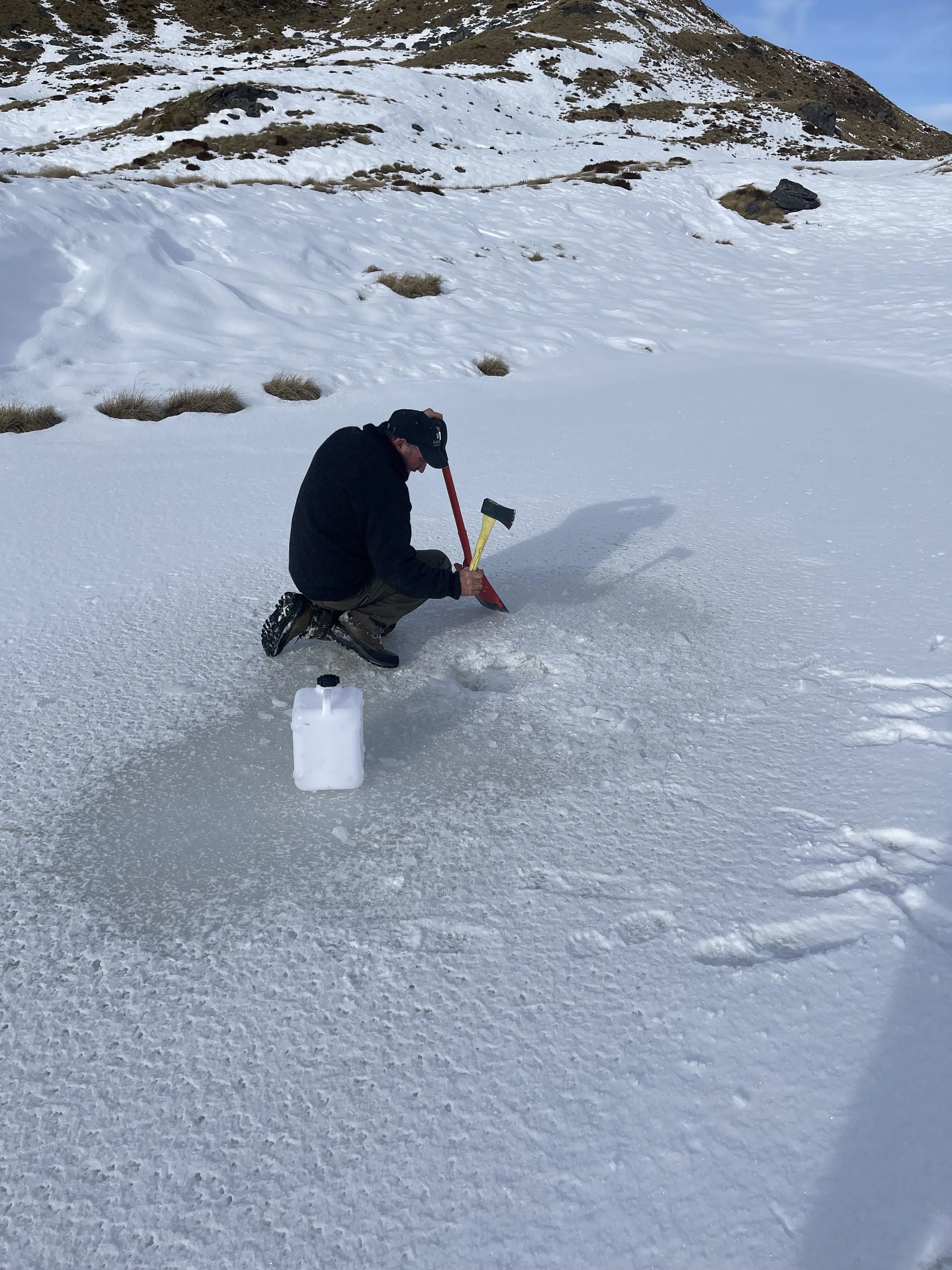 A person kneeling on snow and ice, holding a small axe, next to a plastic container, in a snowy mountainous landscape.
