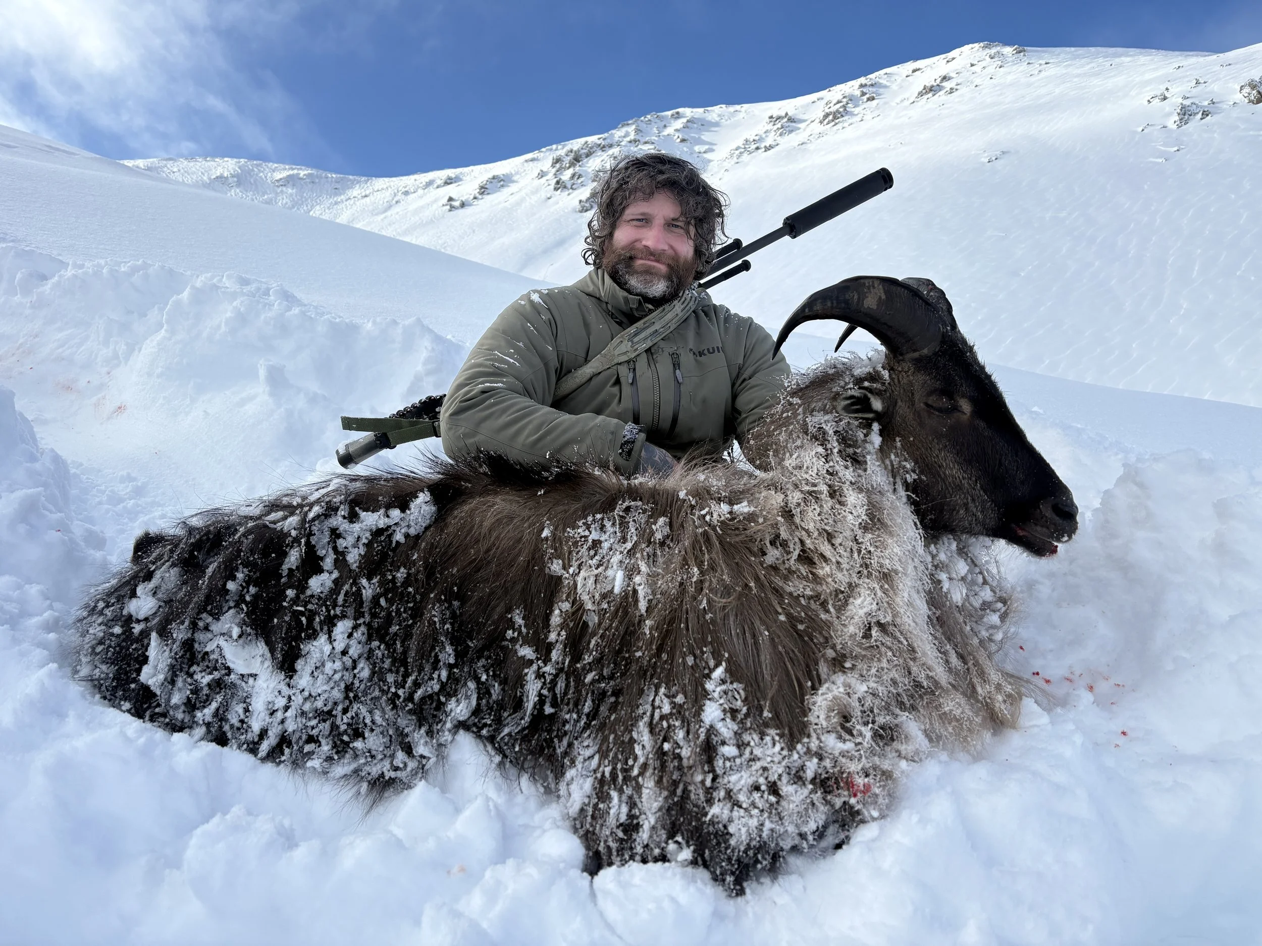 A man with a beard and curly hair kneels in snow holding a deceased mountain Tahr with curved horns; the landscape is snowy with a mountain in the background.