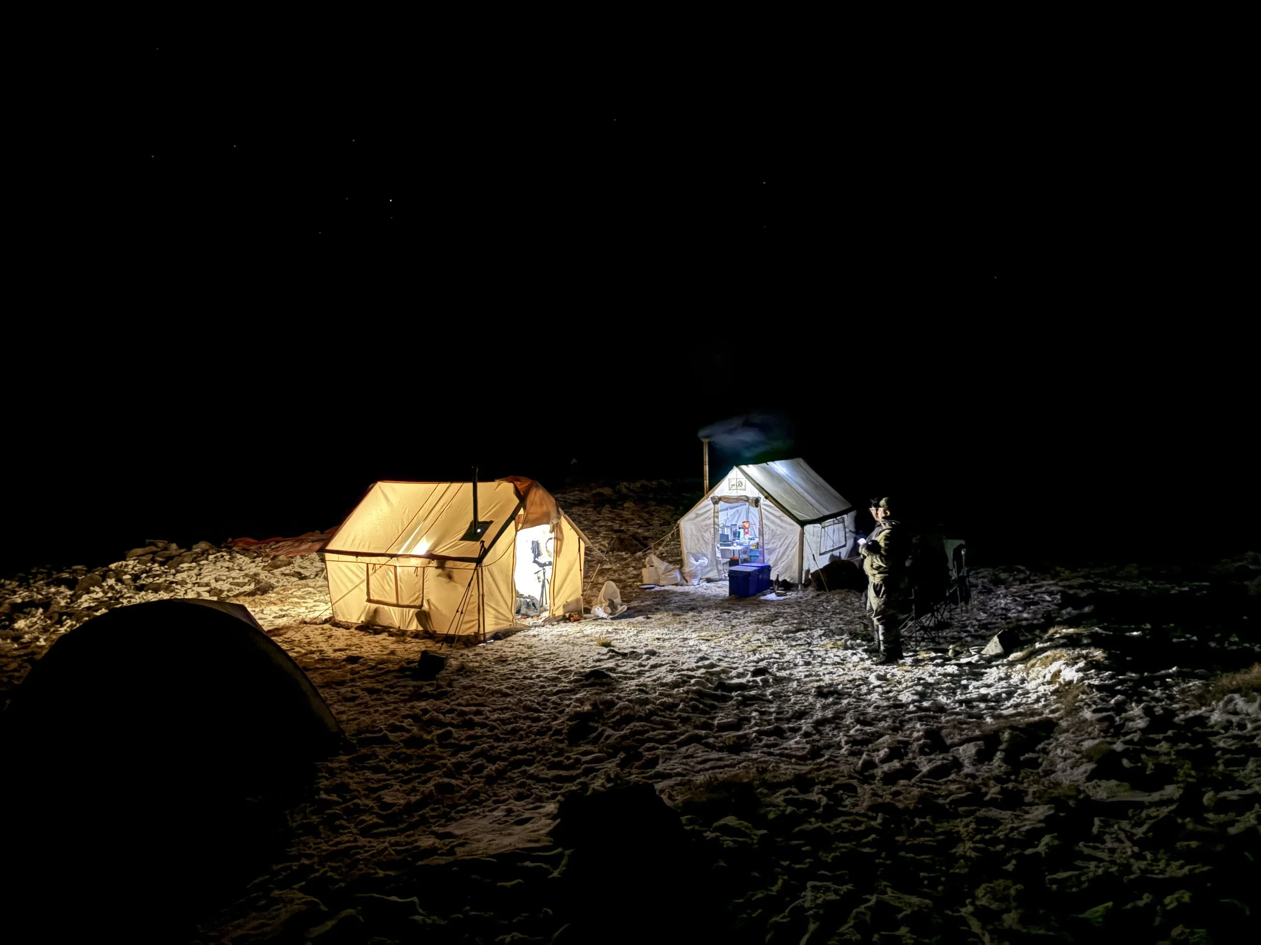 Campers' tents illuminated at night on snowy ground with a person standing nearby, under a starry sky.