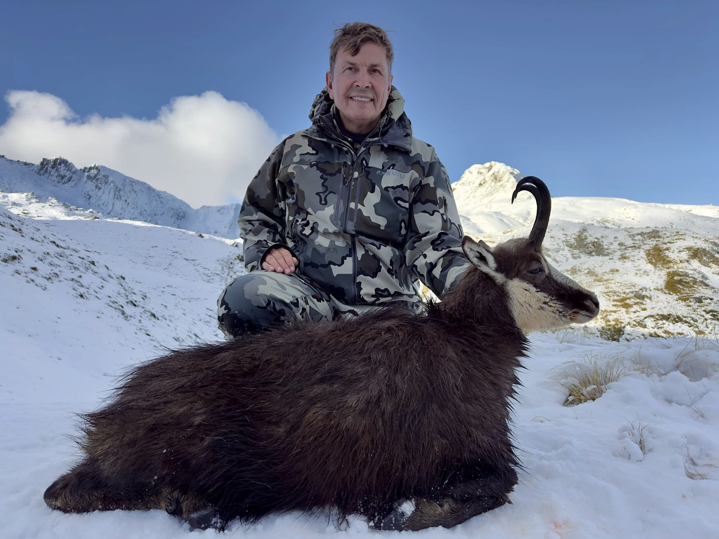 A man in camouflage winter clothing sitting outdoors on snow-covered ground, smiling, with a mountain landscape and blue sky in the background. There is also a llama or alpaca lying in the snow next to him.