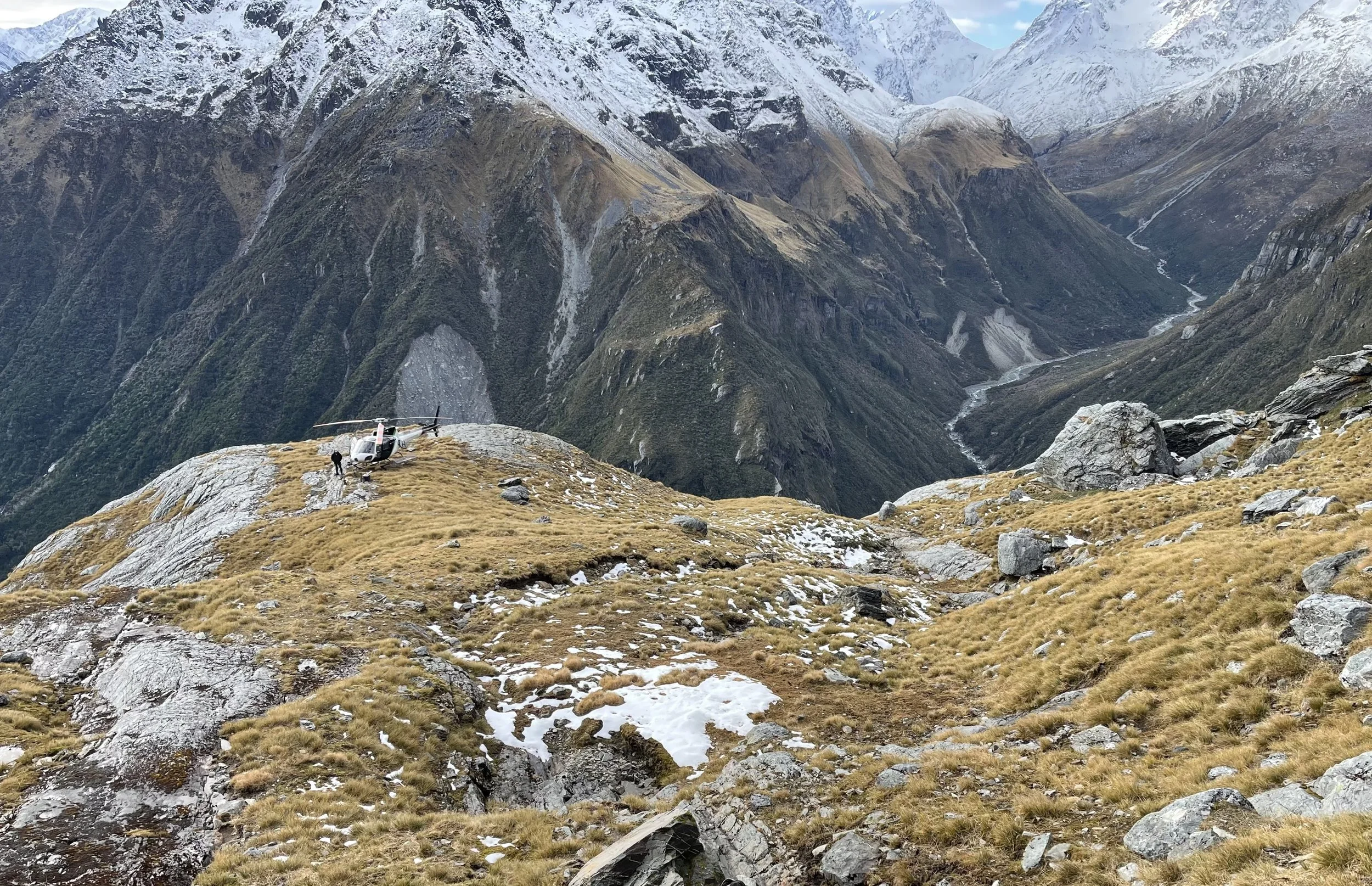 A helicopter parked on a mountain ledge with snow patches and a person standing nearby, surrounded by rugged mountainous terrain with snow-capped peaks in the background.