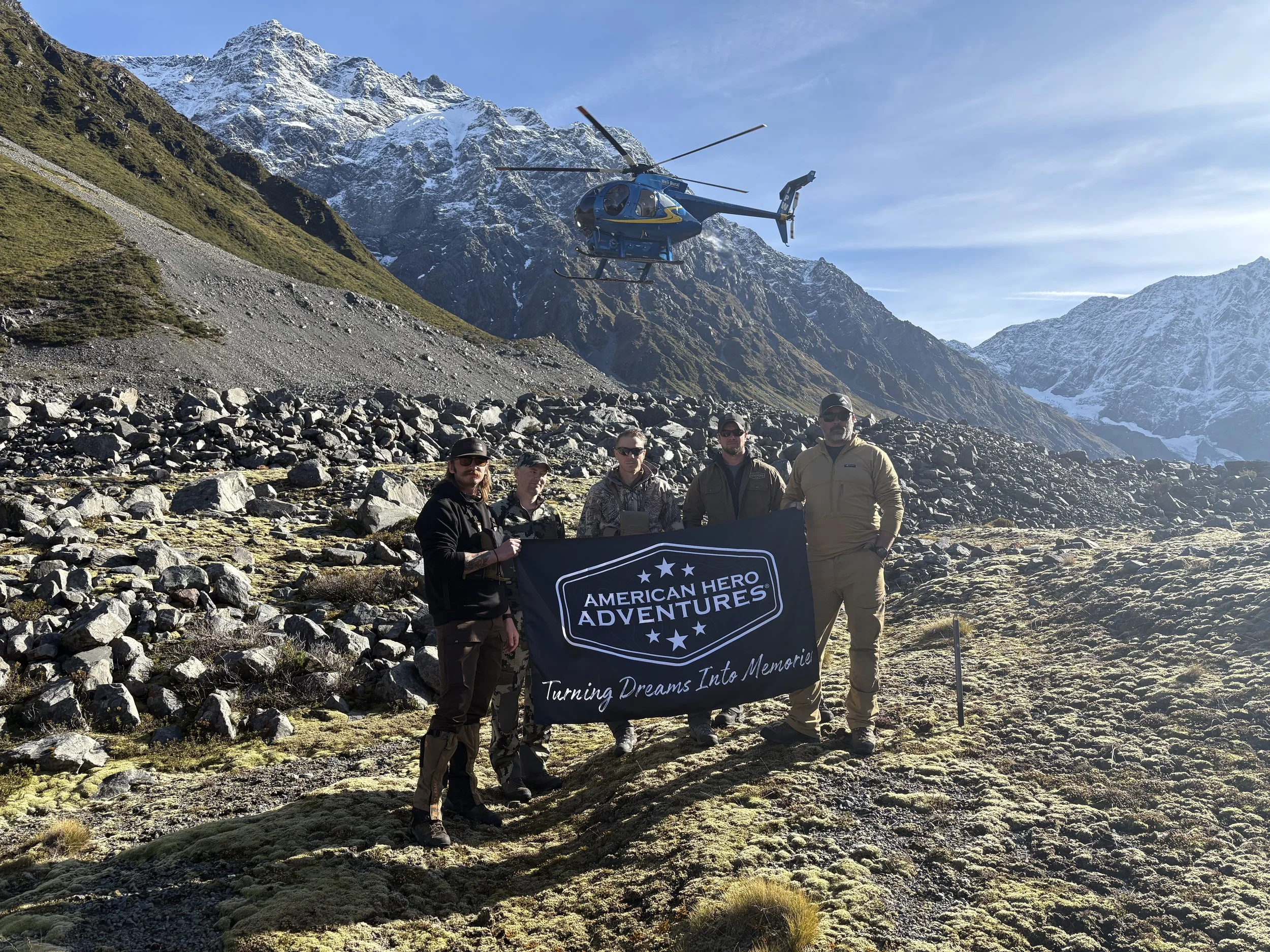 Group of five people holding a black banner with the logo and text of American Hero Adventures, standing in a mountainous landscape with snow-capped peaks, rocky terrain, and a helicopter flying overhead.