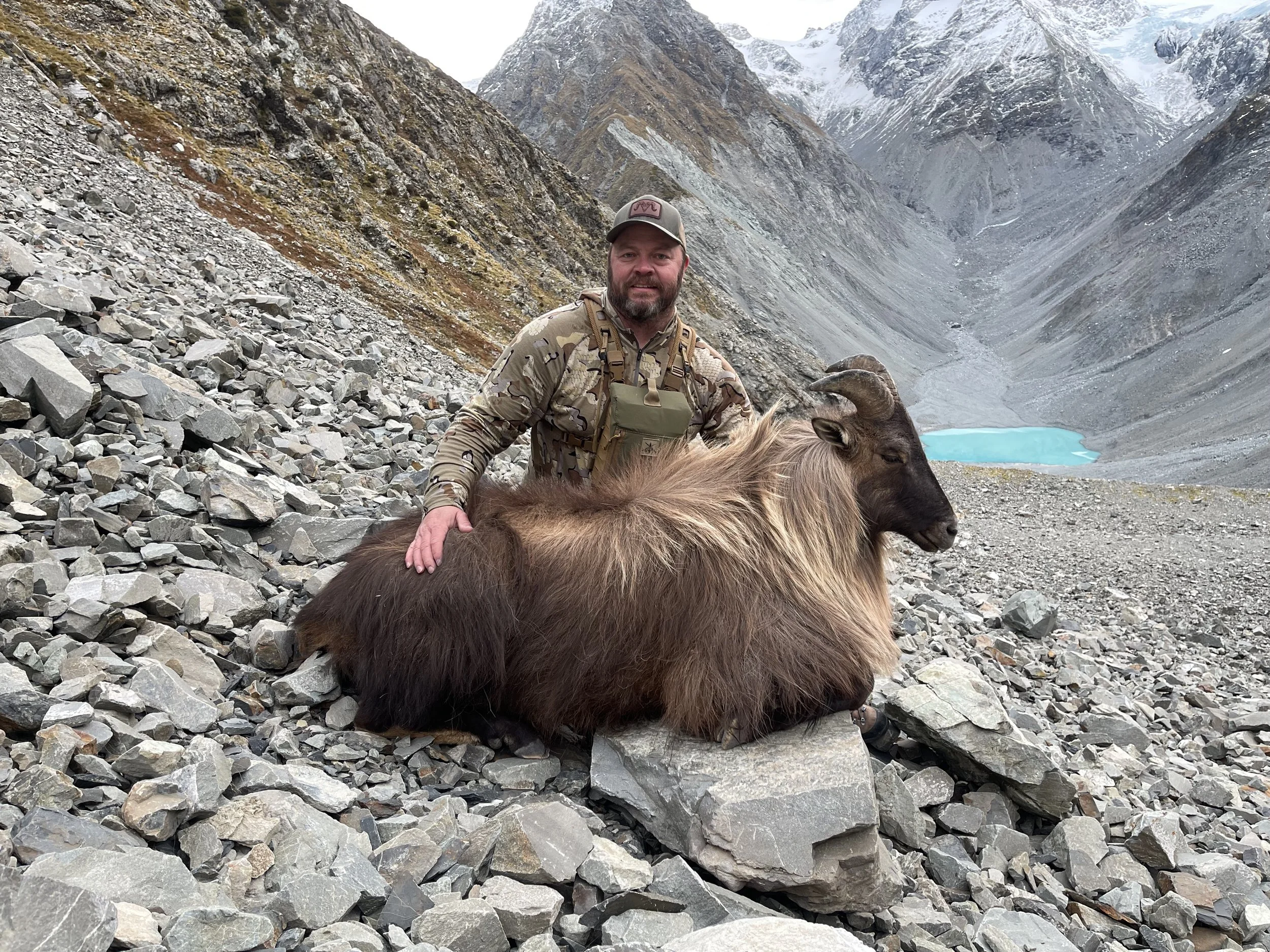 Man in camouflage clothing kneeling with a large tahr on rocky mountain terrain, with a lake and snow-capped peaks in the background. Southern Alps NZ