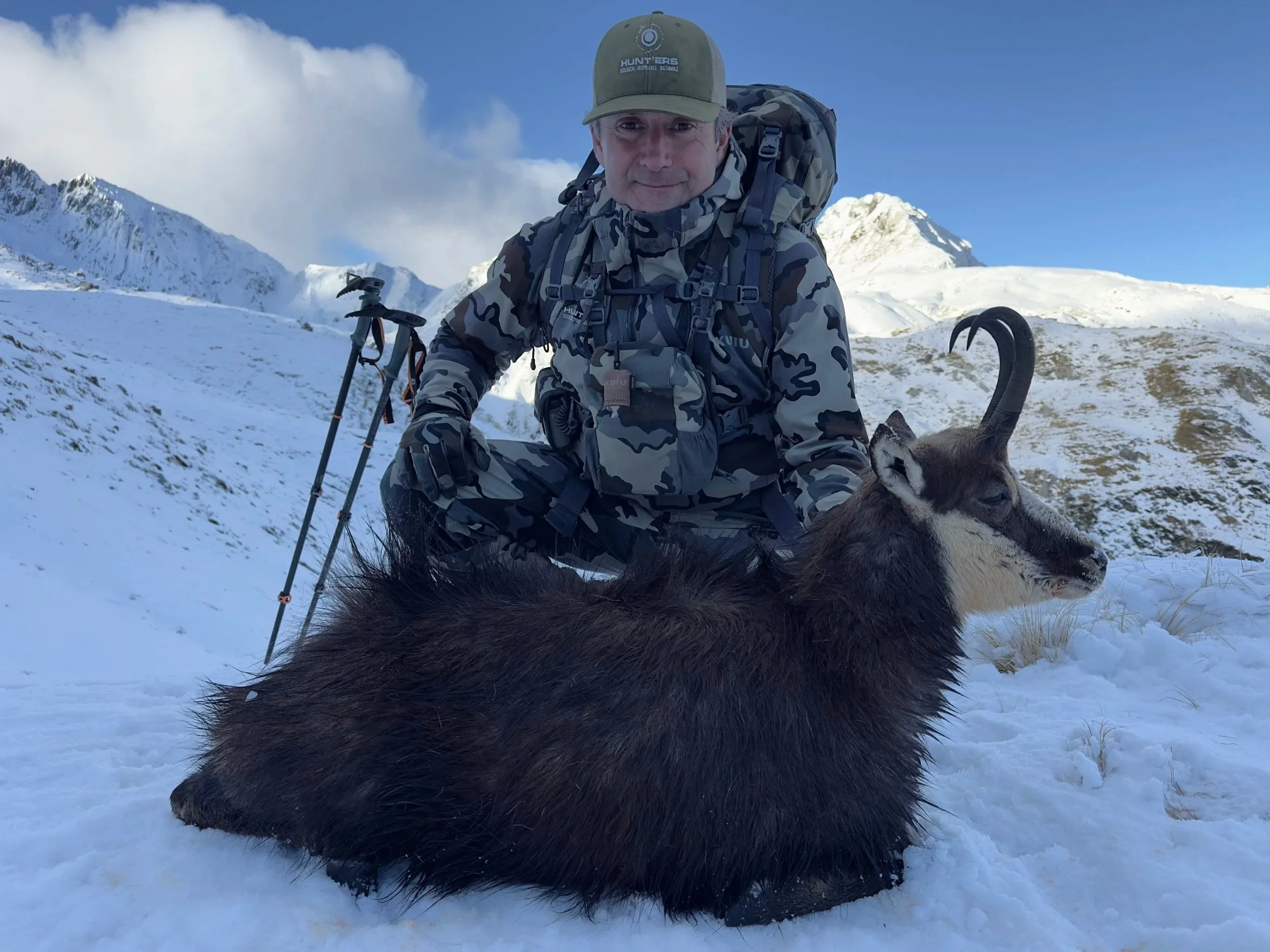 A man dressed in camouflage outdoor gear and a hat, sitting on snow-covered ground with mountains in the background. He has a backpack and trekking poles, and a young goat is lying in front of him.