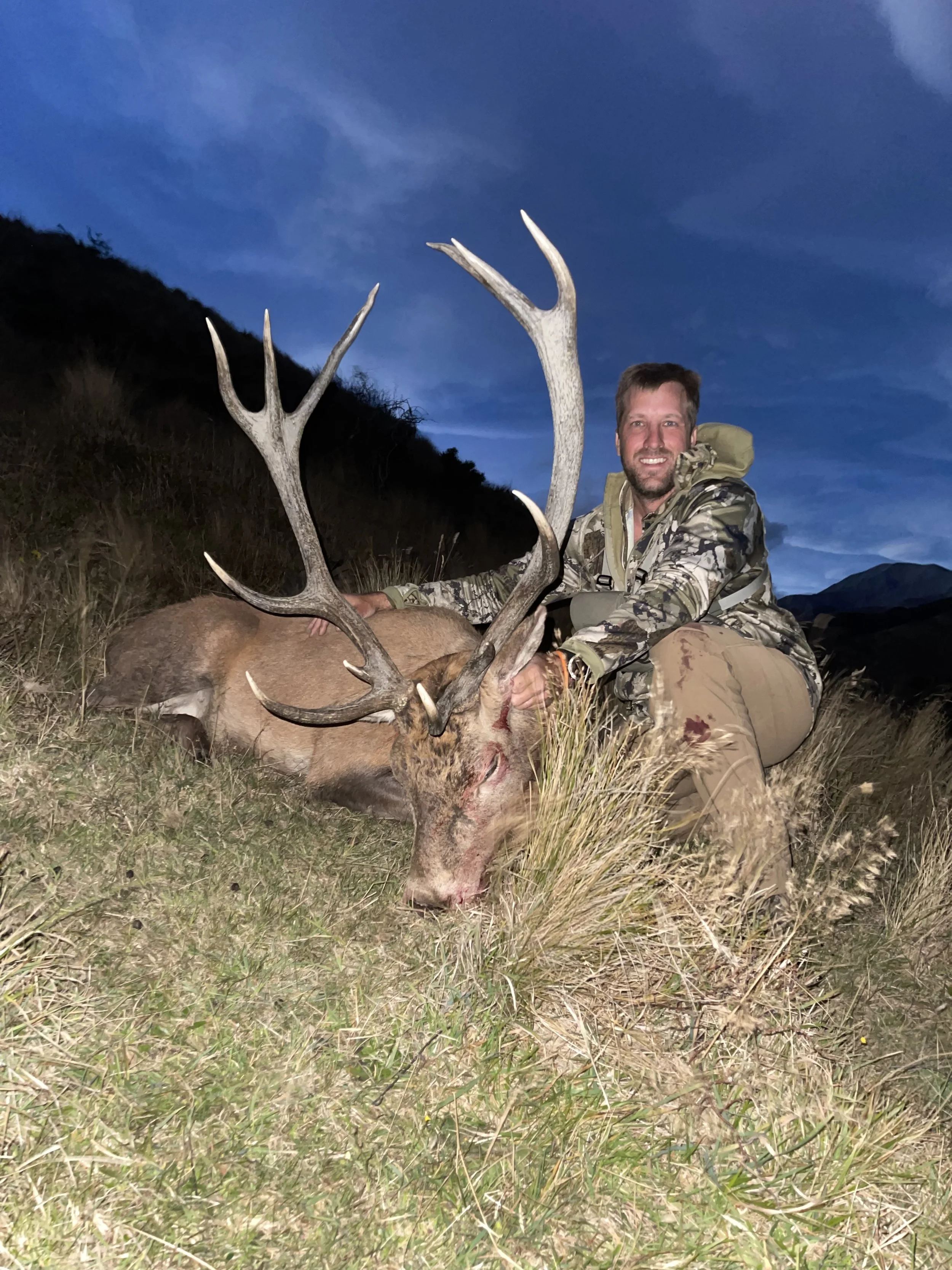 A man in camouflage clothing kneels next to a large stag with impressive antlers, displaying a smile in an outdoor natural setting during dusk or dawn.
