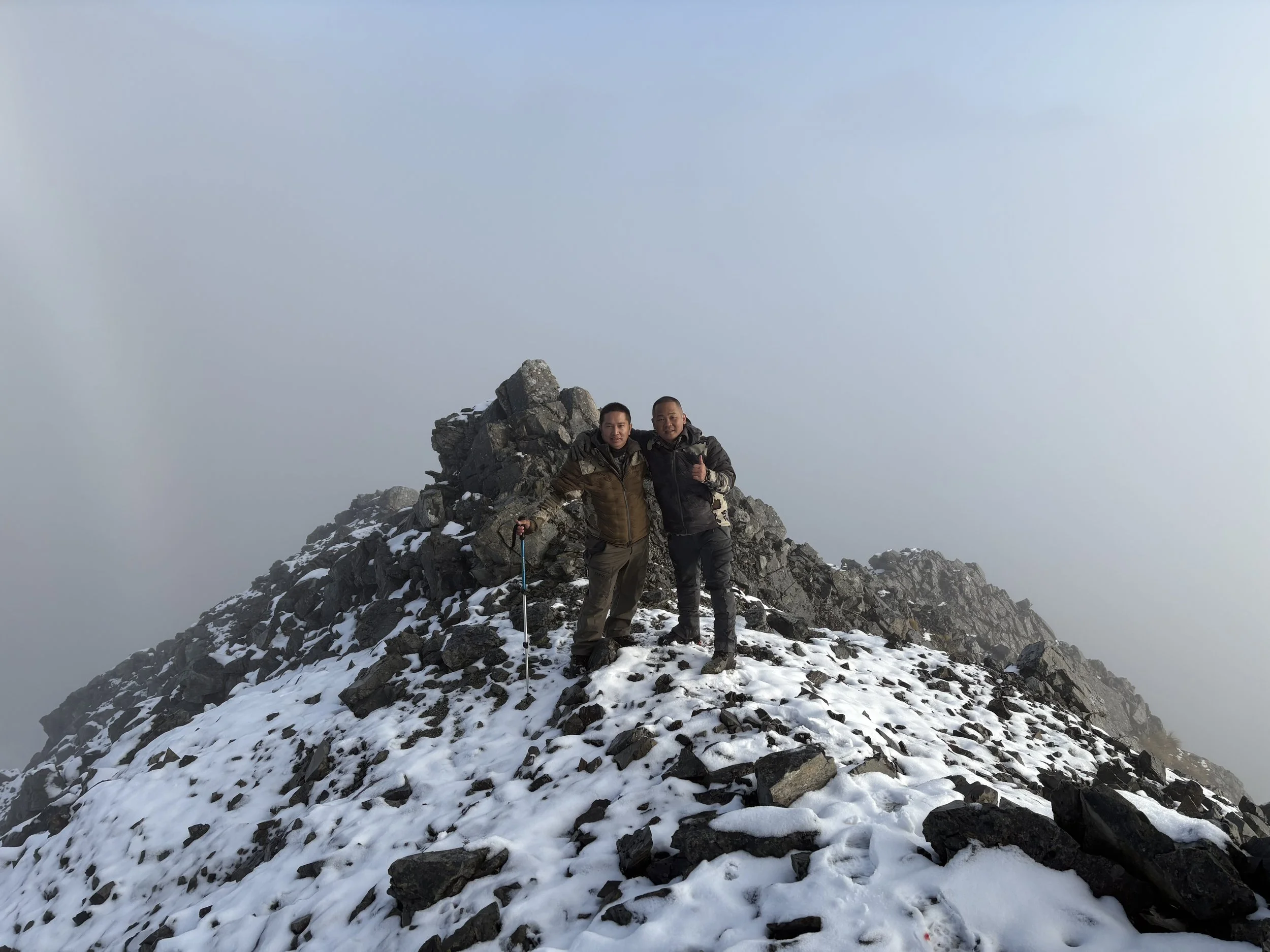 Two men standing on snowy mountain peak with rocky summit and foggy sky in the background, one holding a walking stick, celebrating reaching the top.