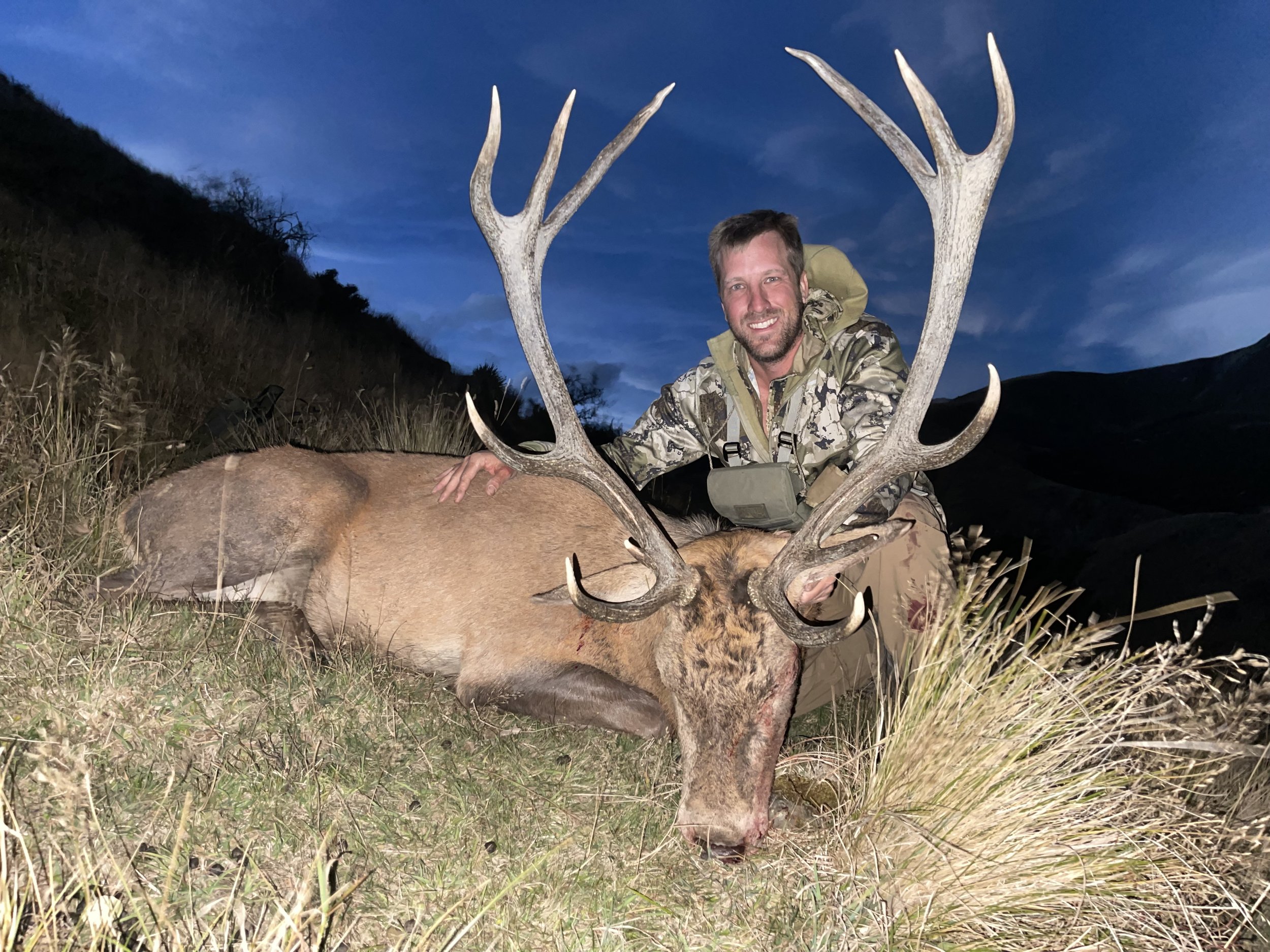 A man wearing camouflage clothing and carrying a backpack, smiling while kneeling next to a large red stag he has hunted, outdoors during dusk or nighttime with a dark sky and hilly landscape in the background.
