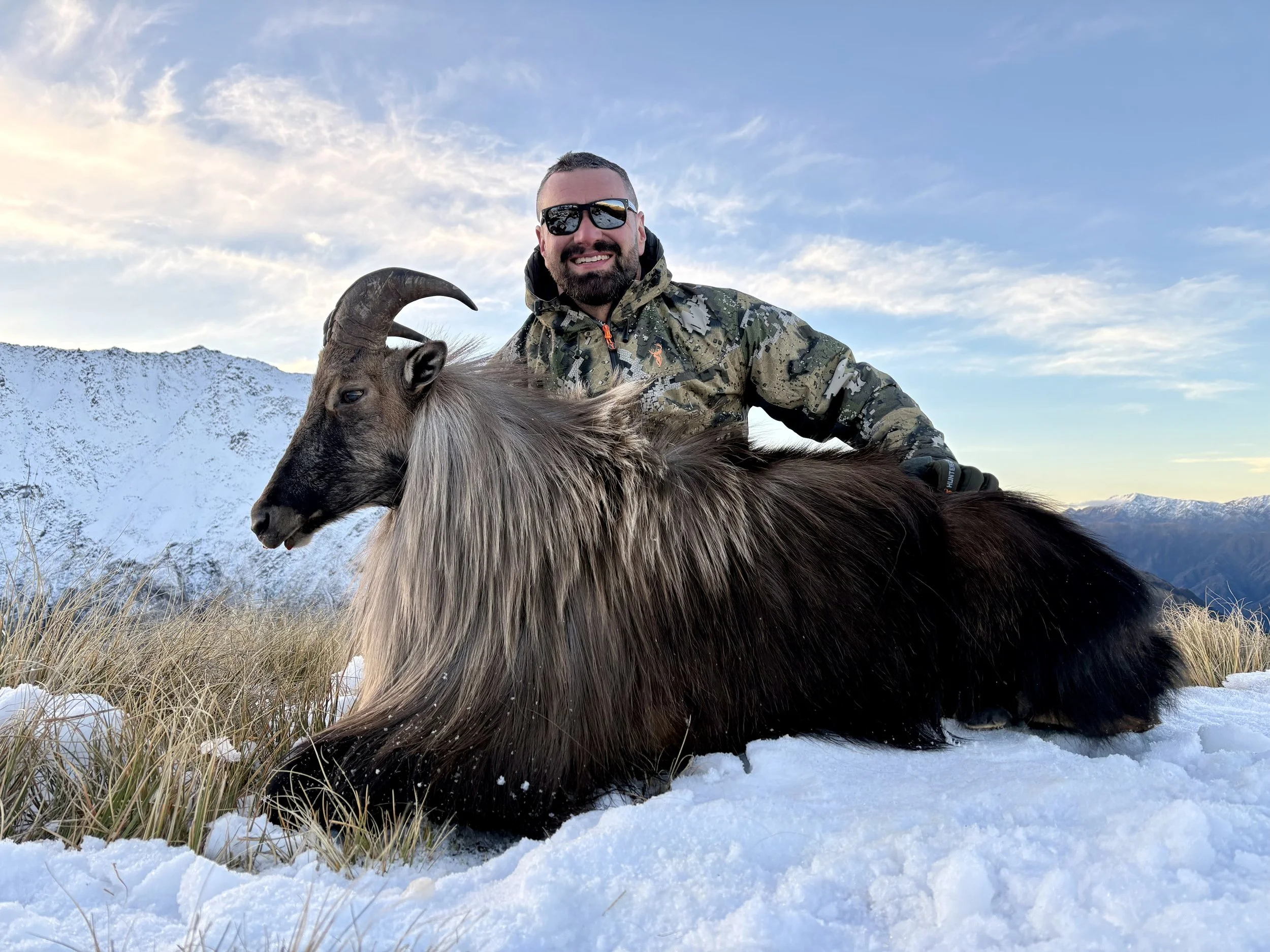 A man wearing sunglasses and camouflage jacket, smiling, sitting on a large brown and black goat in a snowy landscape with mountains and a partly cloudy sky in the background.
