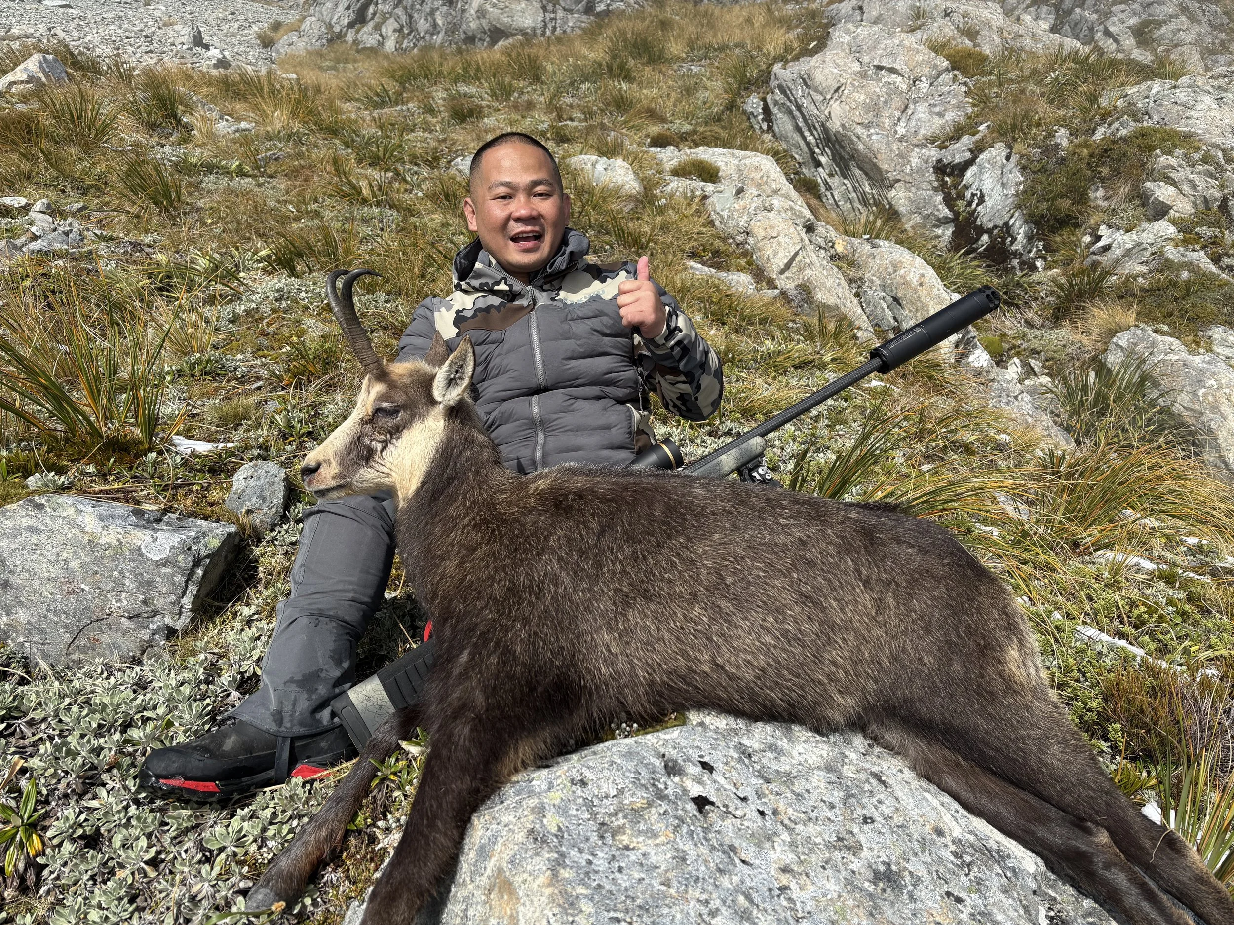 A man sitting on rocky mountain terrain with a rifle, giving a thumbs-up, and resting next to a large mountain goat