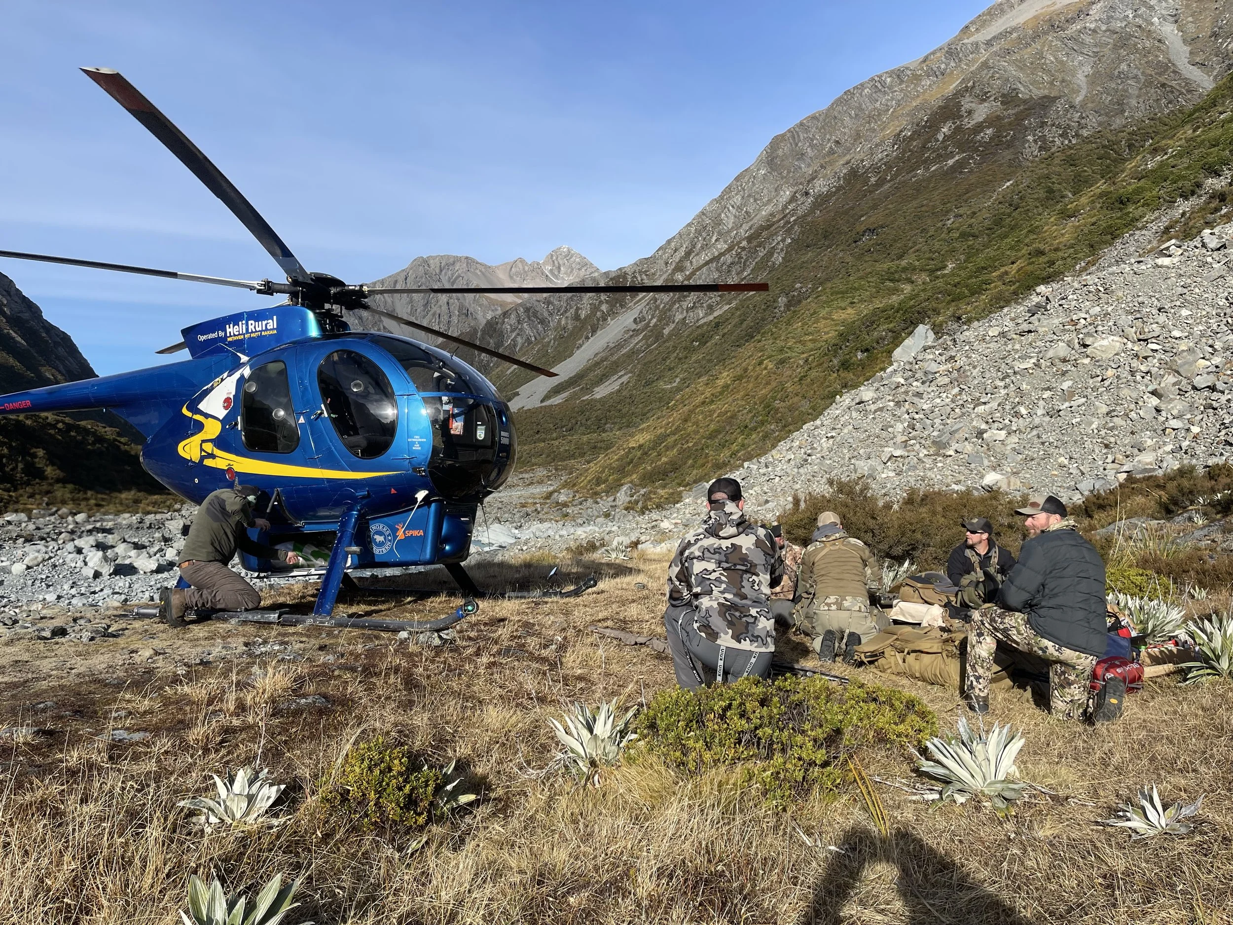 A blue helicopter on a rocky and grassy mountain landscape with a group of people sitting nearby and one person working on the helicopter. they have landed at a remote campsite in the Southern Alps of New Zealand