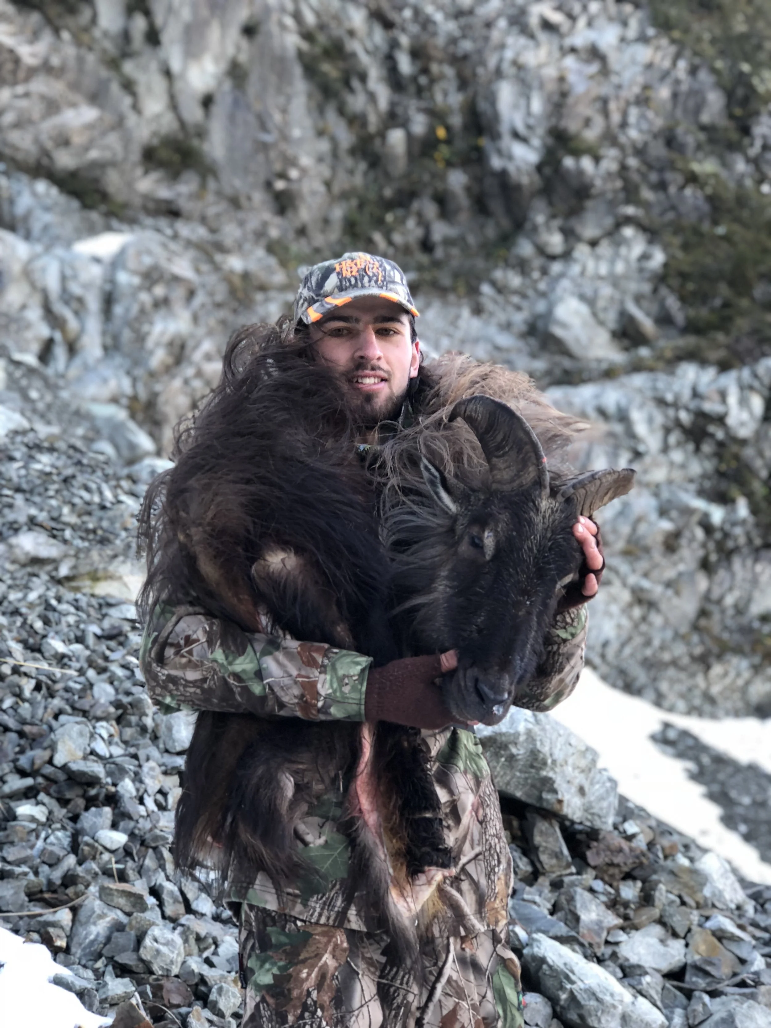 A man in camouflage and a cap holding a bull tahr cape in a rocky mountain landscape.