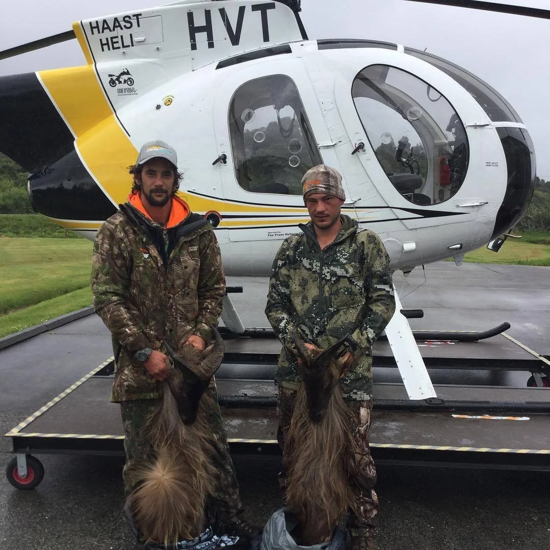 Two hunters in camouflage standing in front of a helicopter holding a hunted animal by its hind legs.