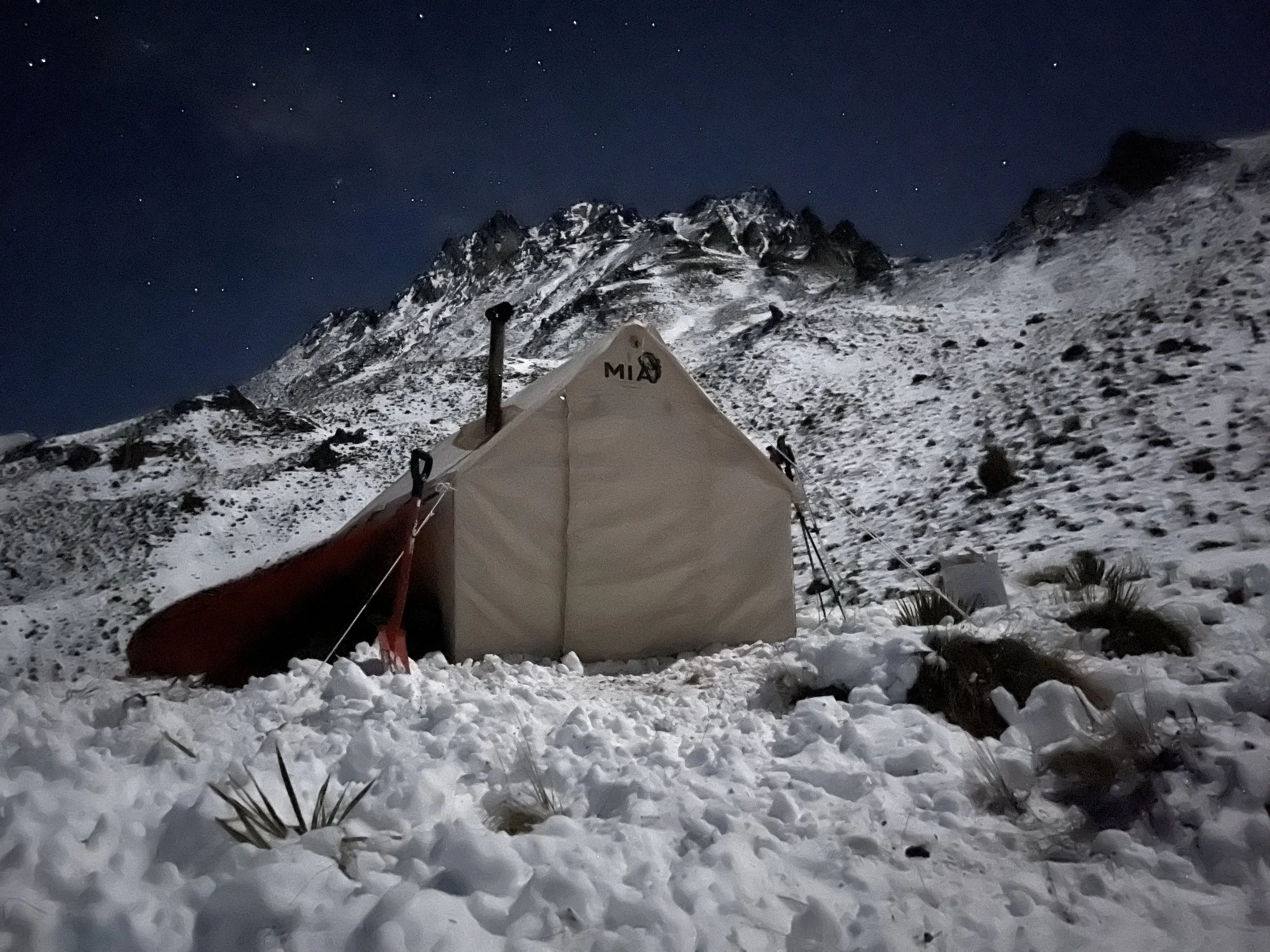 Snow-covered mountain landscape at night with a tent and a kayak in the foreground.