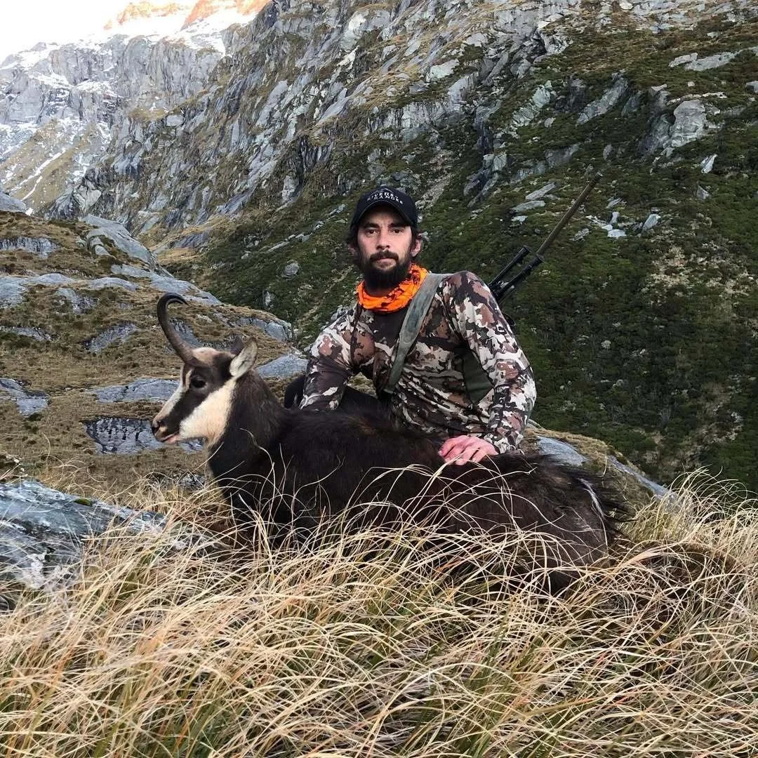 A man dressed in camouflage clothing with a hunting rifle on his back, sitting on rocky terrain amidst tall dry grass, with a mountain and green hillside in the background. He is petting a goat.