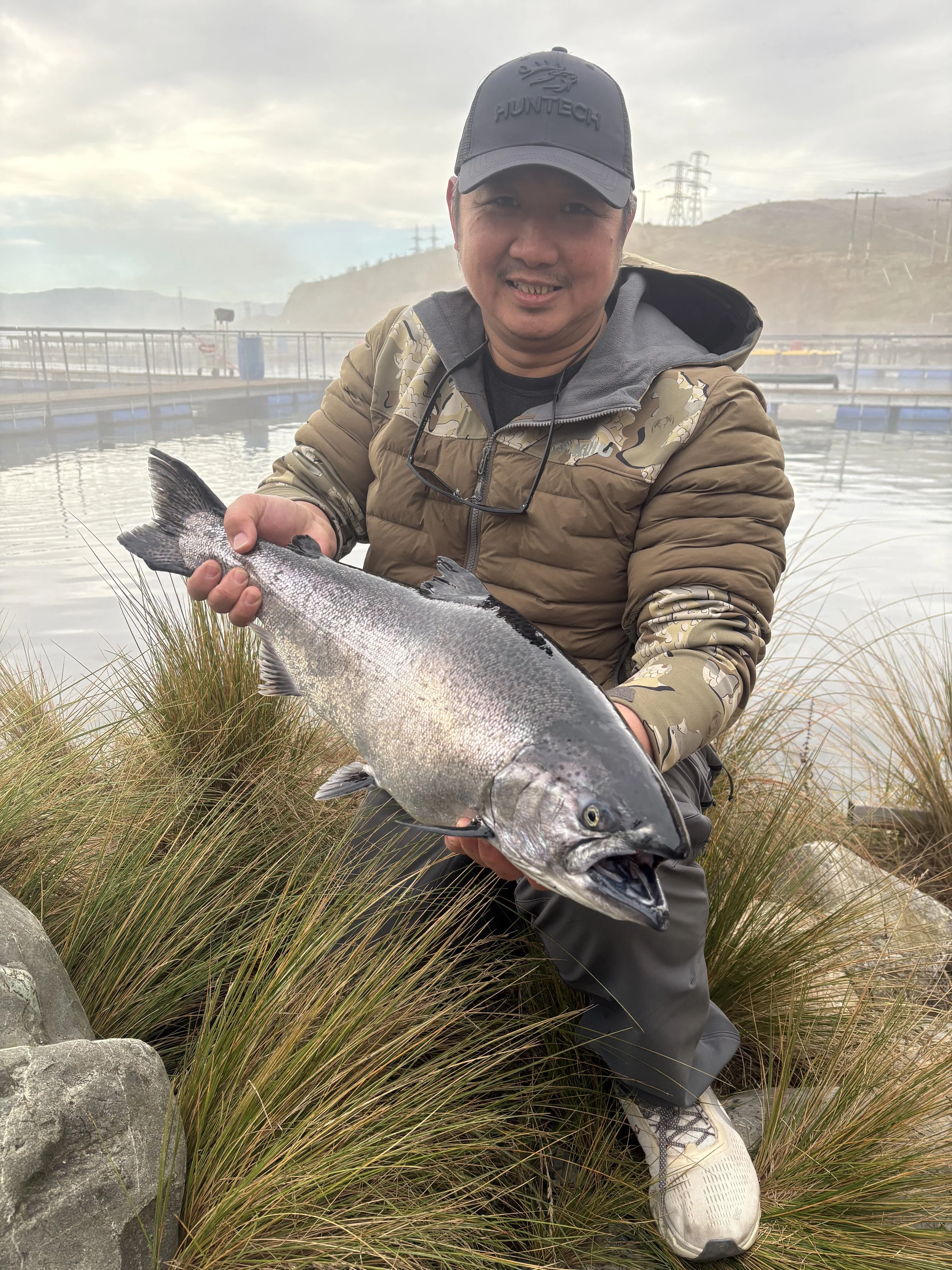 Man smiling and holding a large fish in an outdoor fishing area with water and hills in the background.