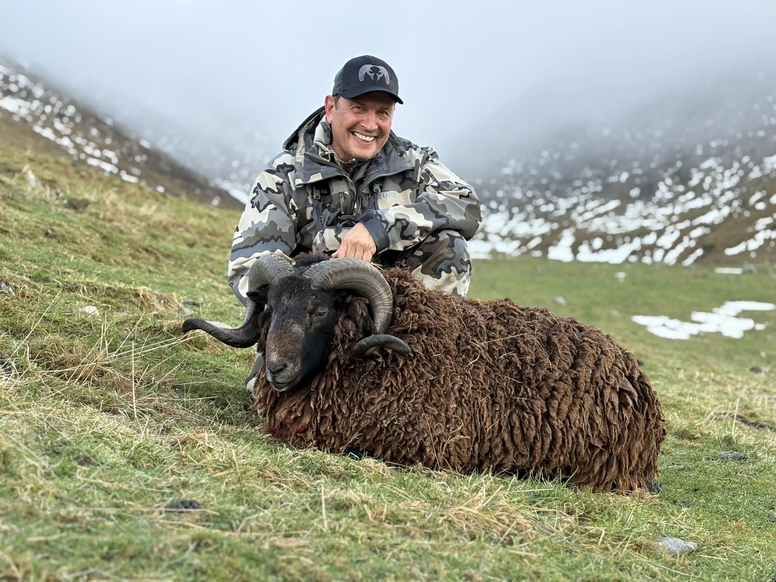 Man smiling in camouflage jacket squatting next to a large, woolly black sheep, called an arapawa ram, with curved horns in a grassy, mountainous landscape with patches of snow and cloudy sky.