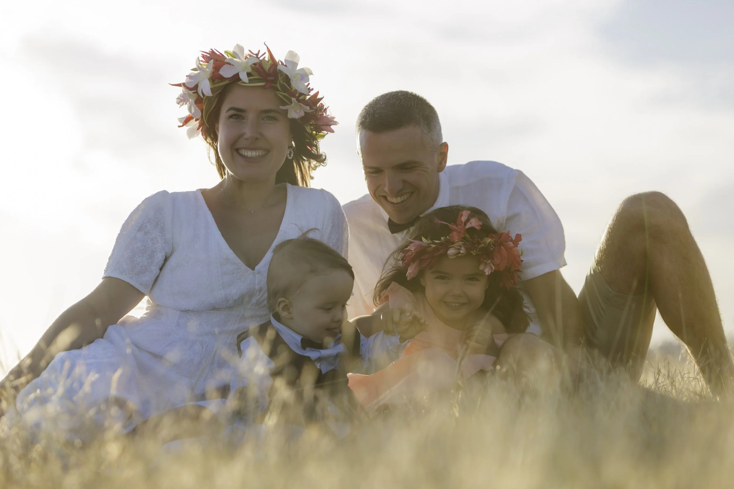 Une famille souriante portant des couronnes de fleurs, assise dans un champ lors d'une séance photo en plein air au coucher du soleil.
