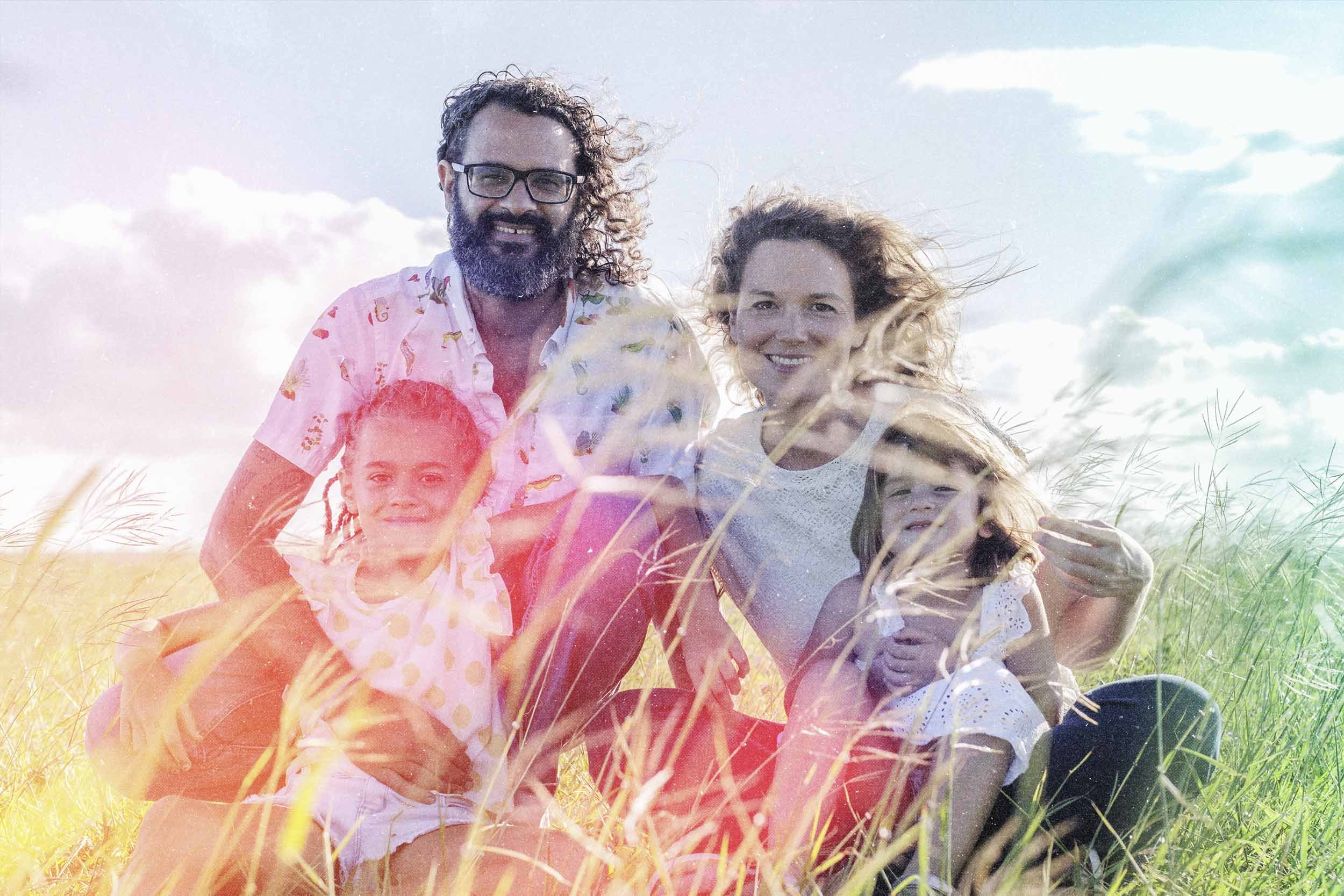 Une famille de quatre personnes souriantes assises dans un champ d'herbe verte, sous un ciel partiellement nuageux, en plein air lors d'une journée ensoleillée.