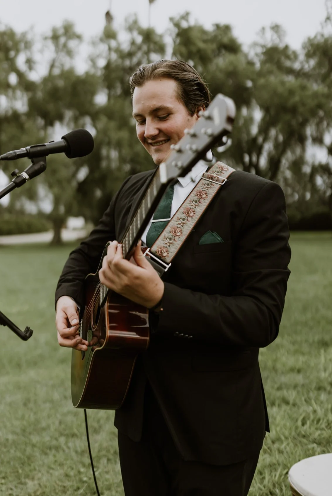 Young man in a black suit playing an acoustic guitar outdoors near a microphone, smiling with trees in the background.