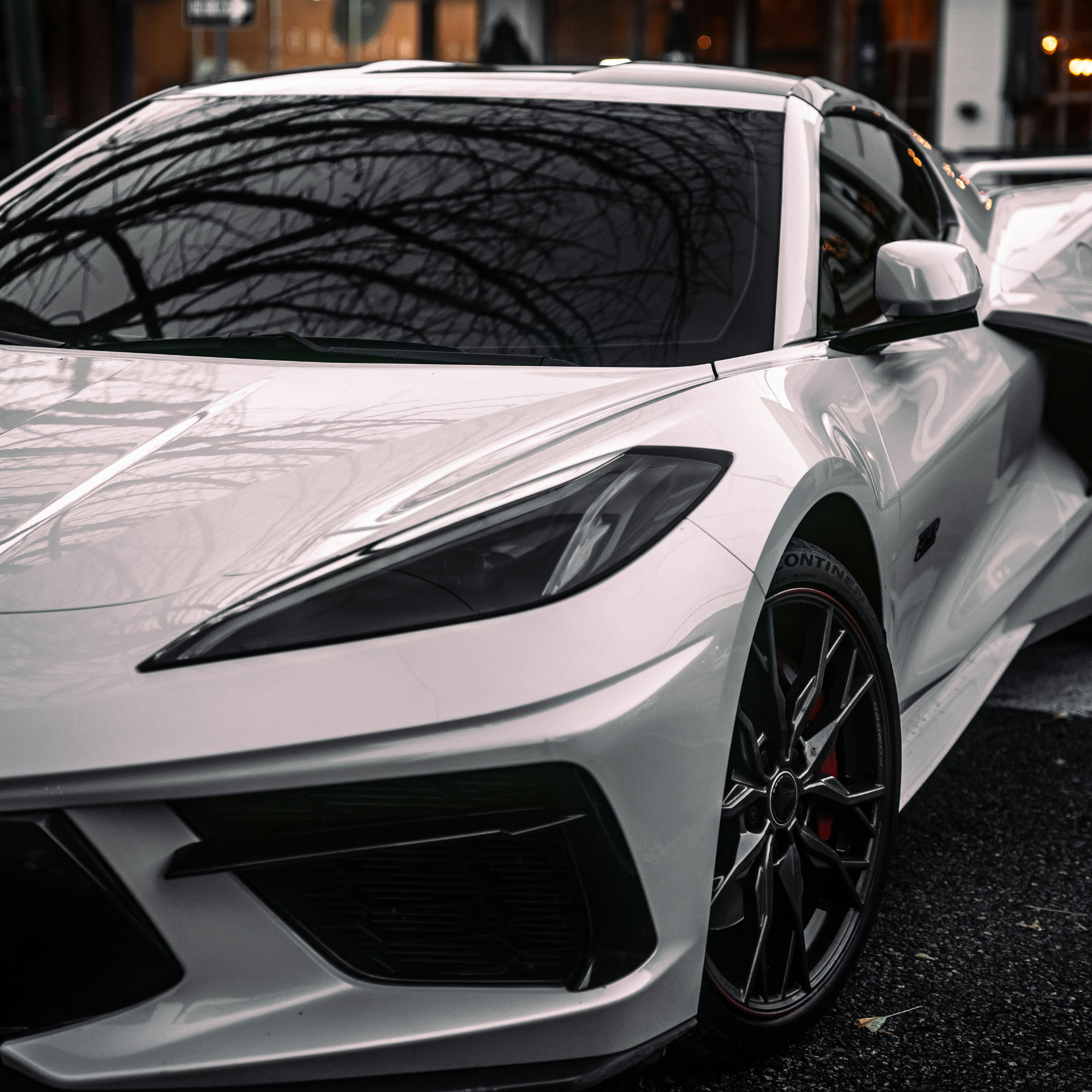 Close-up of a luxury sports car with a sleek silver exterior, sharply designed headlight, and aerodynamic features parked outdoors, with trees casting reflections on its tinted windshield.