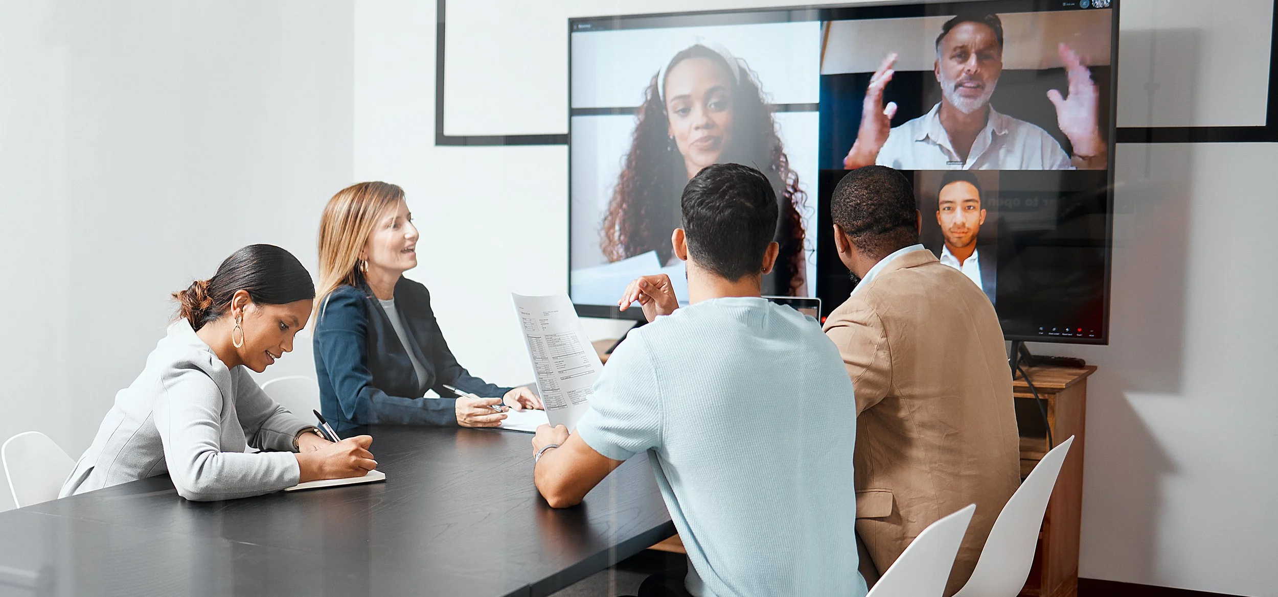 a corporate conference room with four individuals is interacting with colleagues on a large screen video call. The speaker has his hands animatedly in suspension.