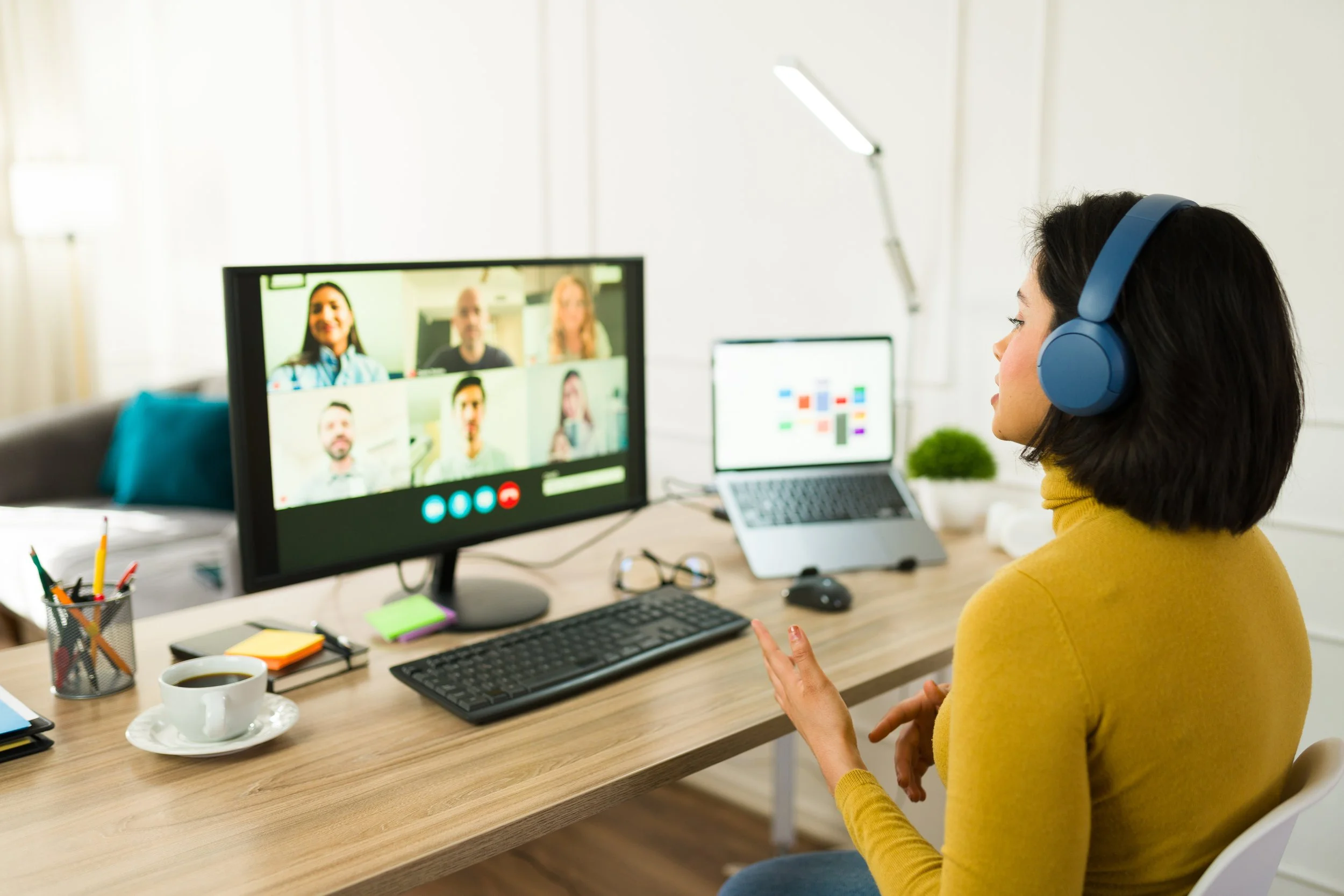 person in mustard colored turtle neck is speaking on a video call to a group of people. They have two screens in front of them and are wearing headphones.