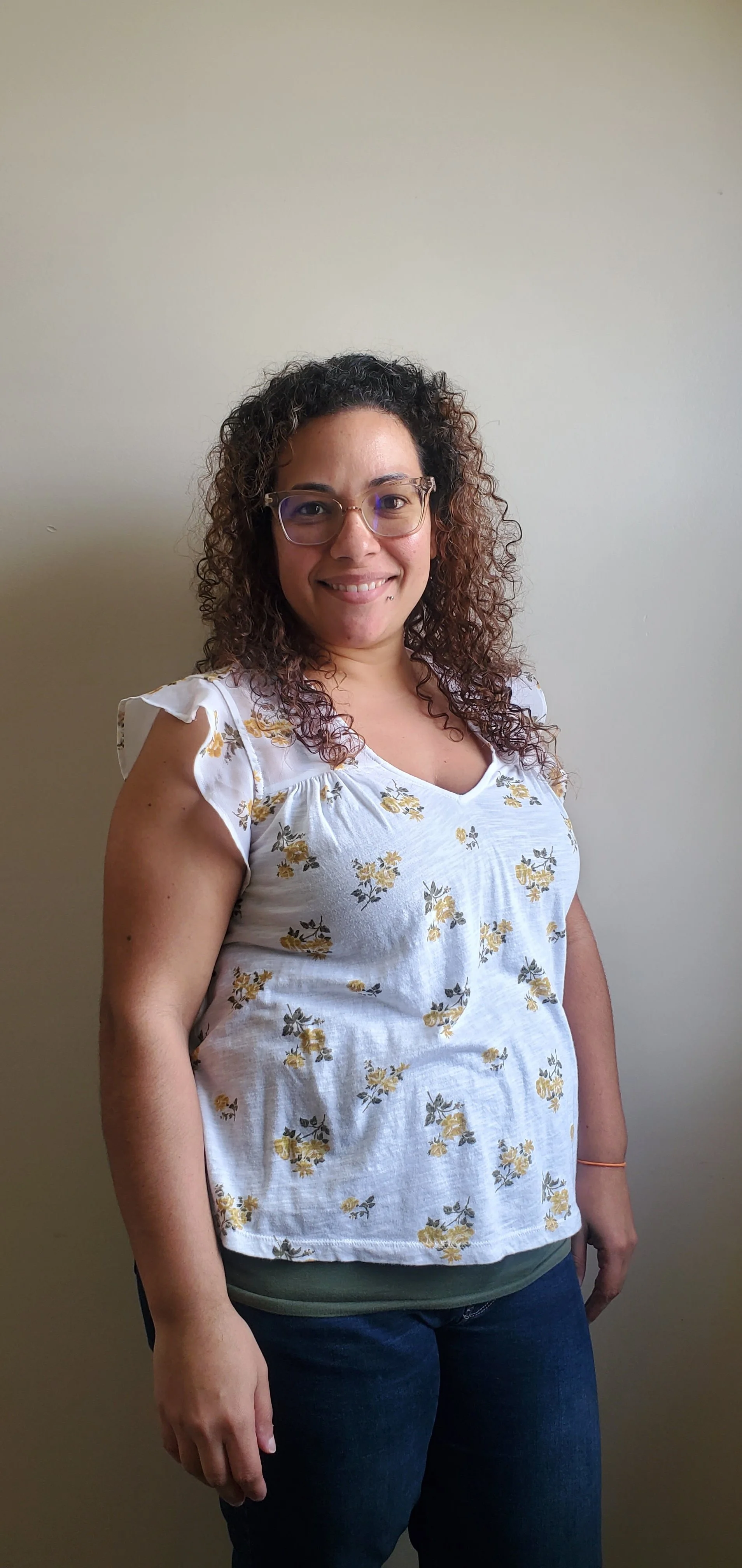 A woman with curly hair wearing glasses, a white floral top, and dark jeans, standing against a plain light-colored wall and smiling at the camera.