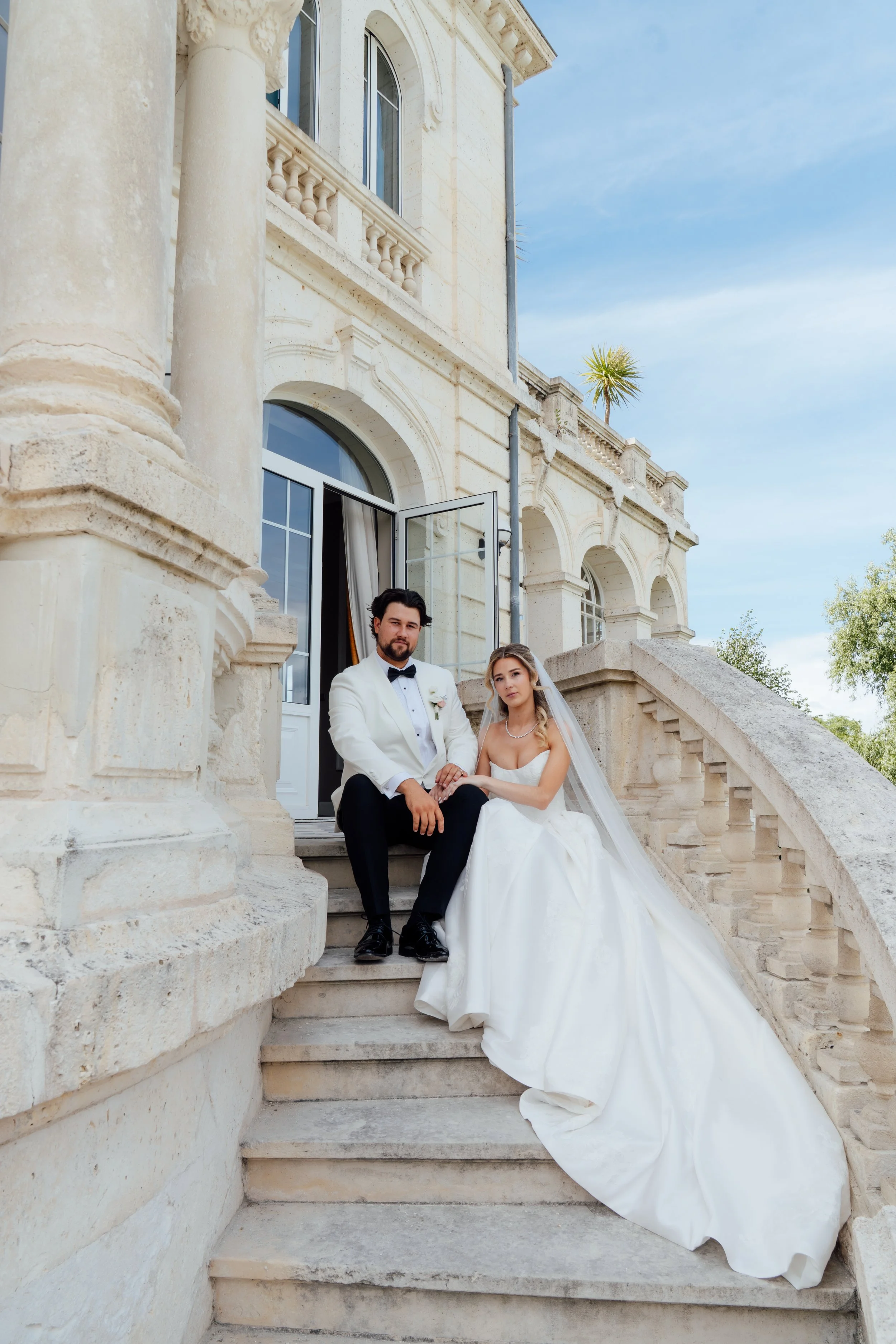 A newly married couple sitting on stone stairs outside a large, elegant building with arched windows and classical architectural details. The groom is in a white tuxedo with a black bow tie, and the bride is in a white wedding gown with a long train, holding hands and looking at the camera.