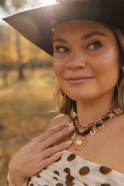 A woman wearing a black wide-brimmed hat and jewelry, with a blurred autumn background.