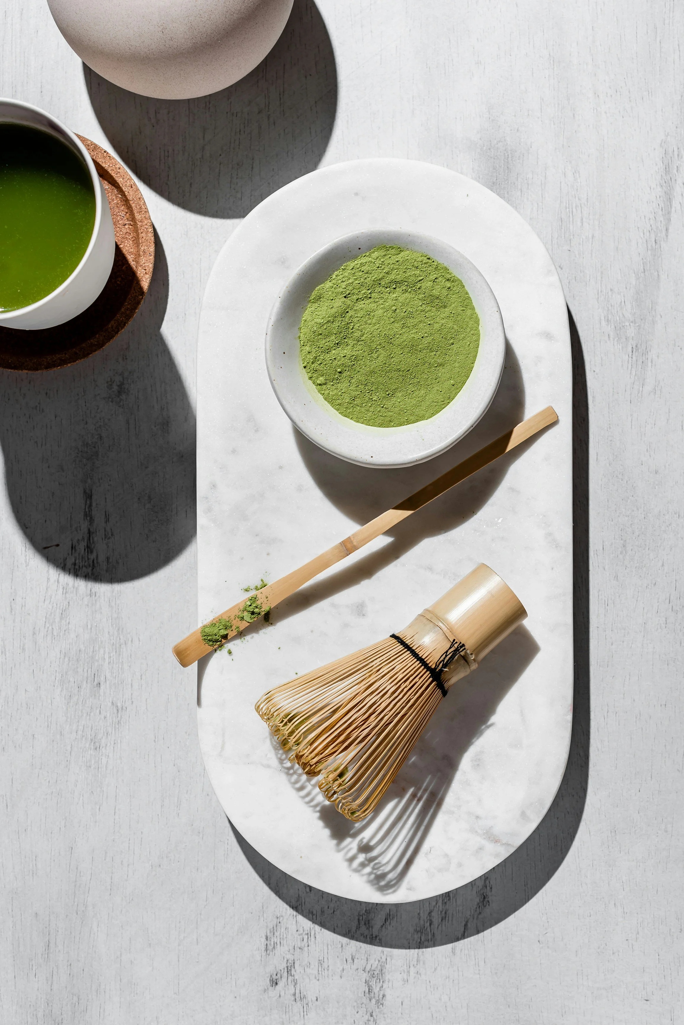 A white oval platter holding matcha a type of Japanese green tea, a bamboo whisk, and a bamboo scoop, with a cup of prepared matcha tea and a white ceramic jar on a light gray surface.