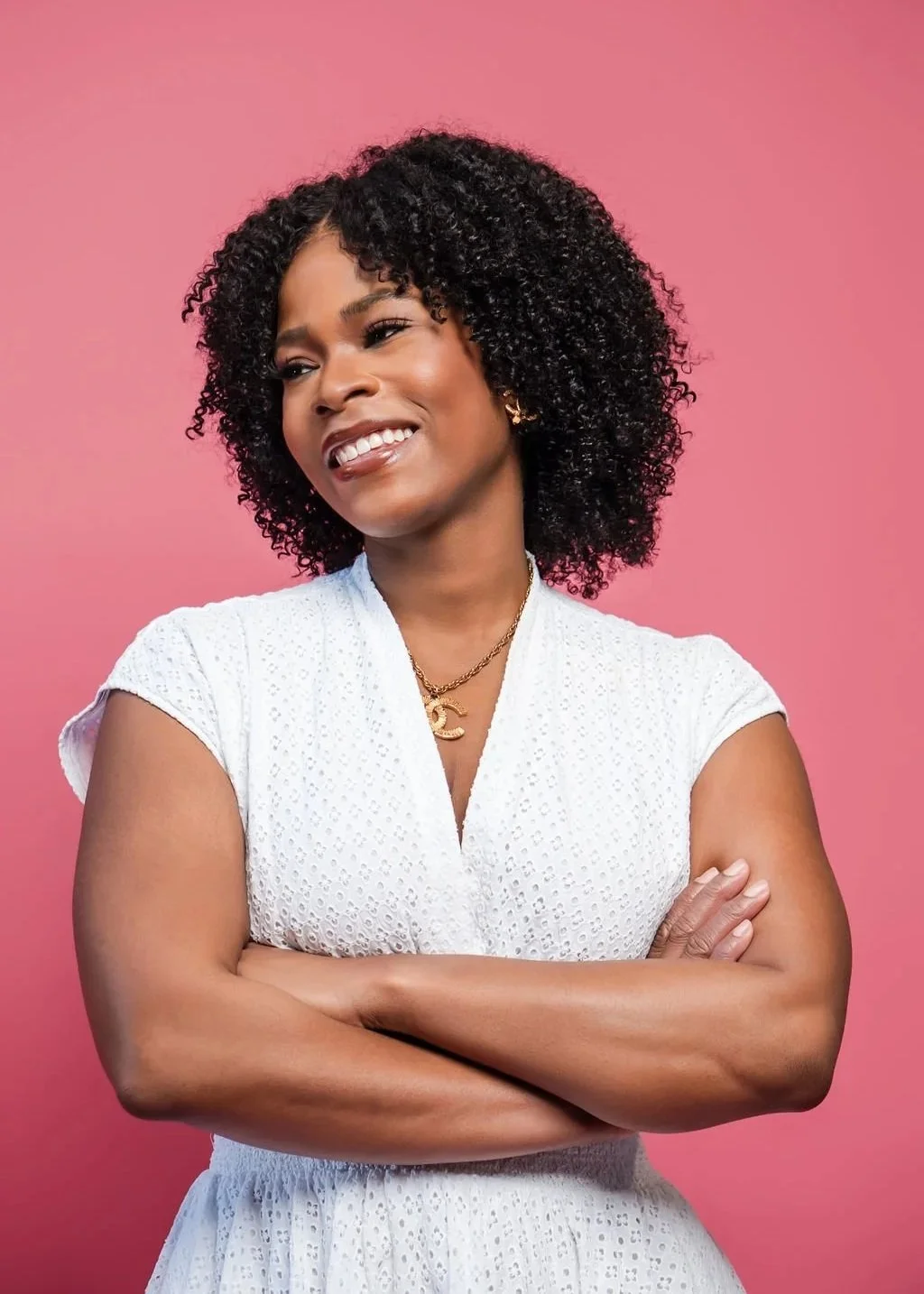 A woman with curly black hair, smiling with arms crossed against a pink background, wearing a white eyelet dress and gold jewelry.