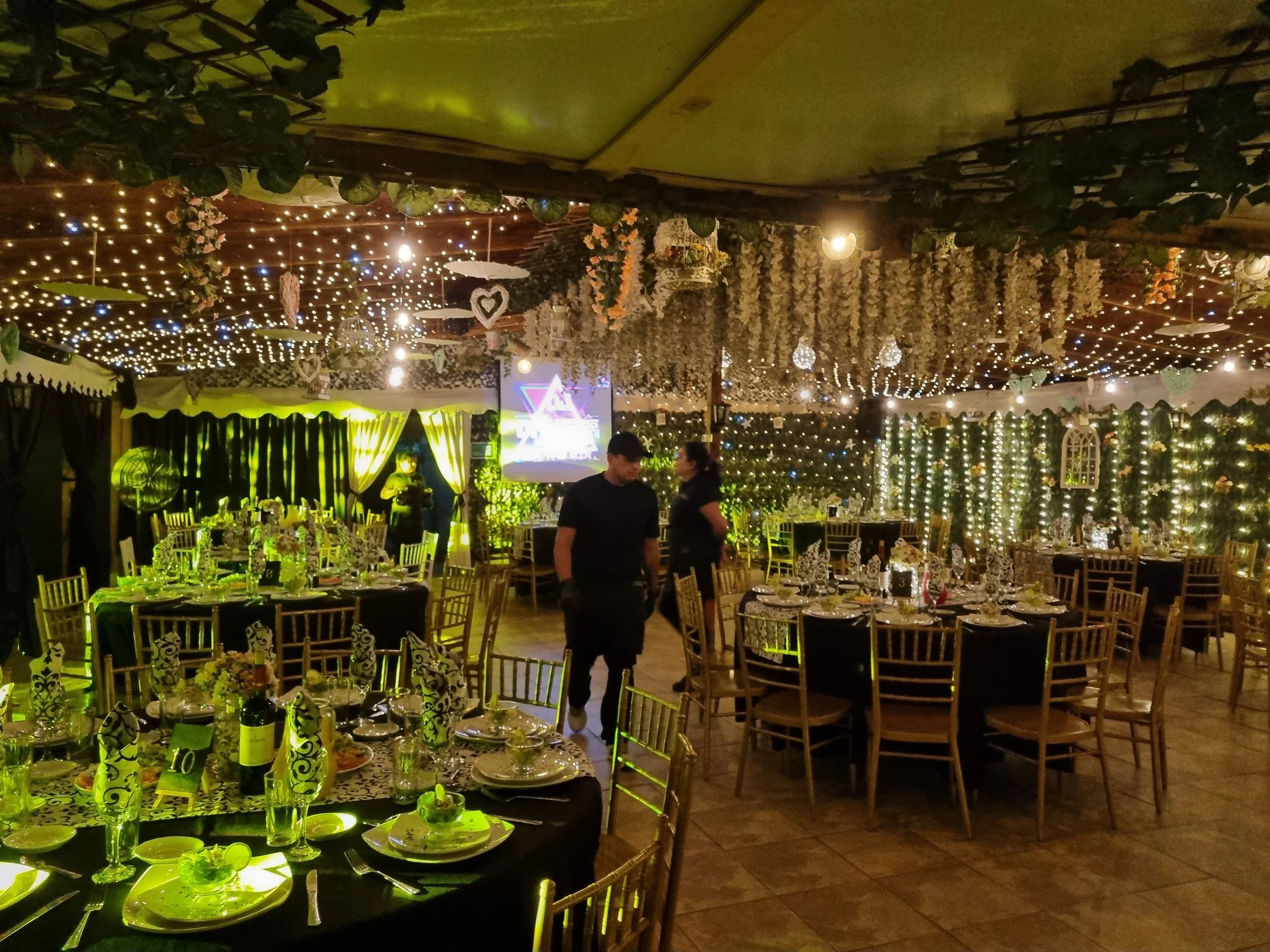 Salón decorado con luces y guirnaldas para una celebración, mesas vestidas para una comida especial, y dos personas conversando en el centro.