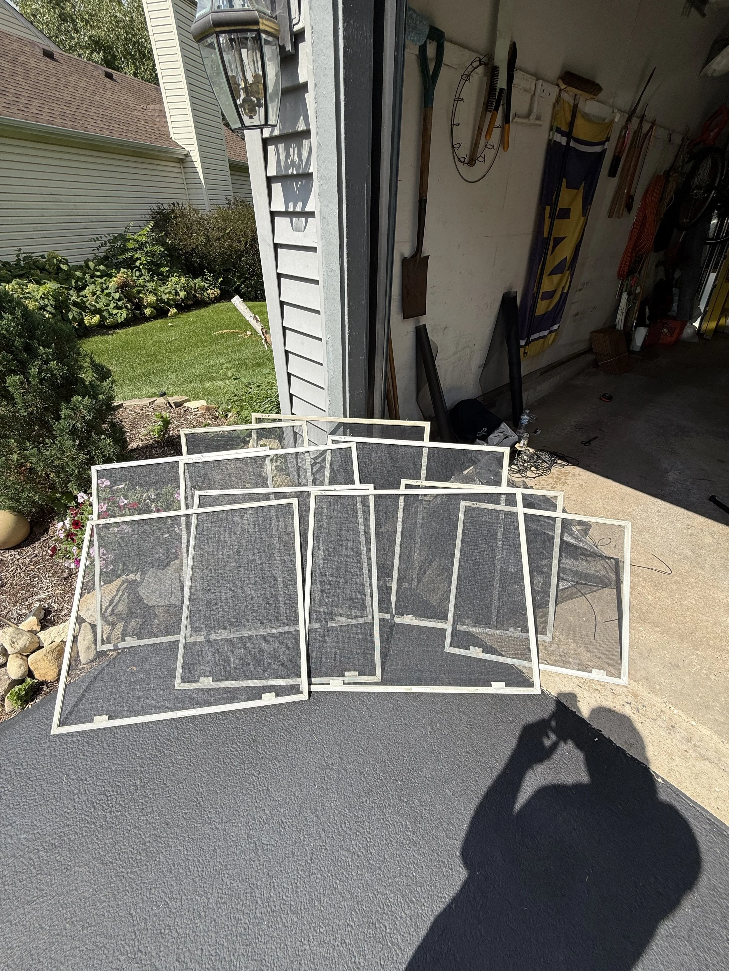 Eight window screens leaning against a garage wall outside, with a garden and house in the background.