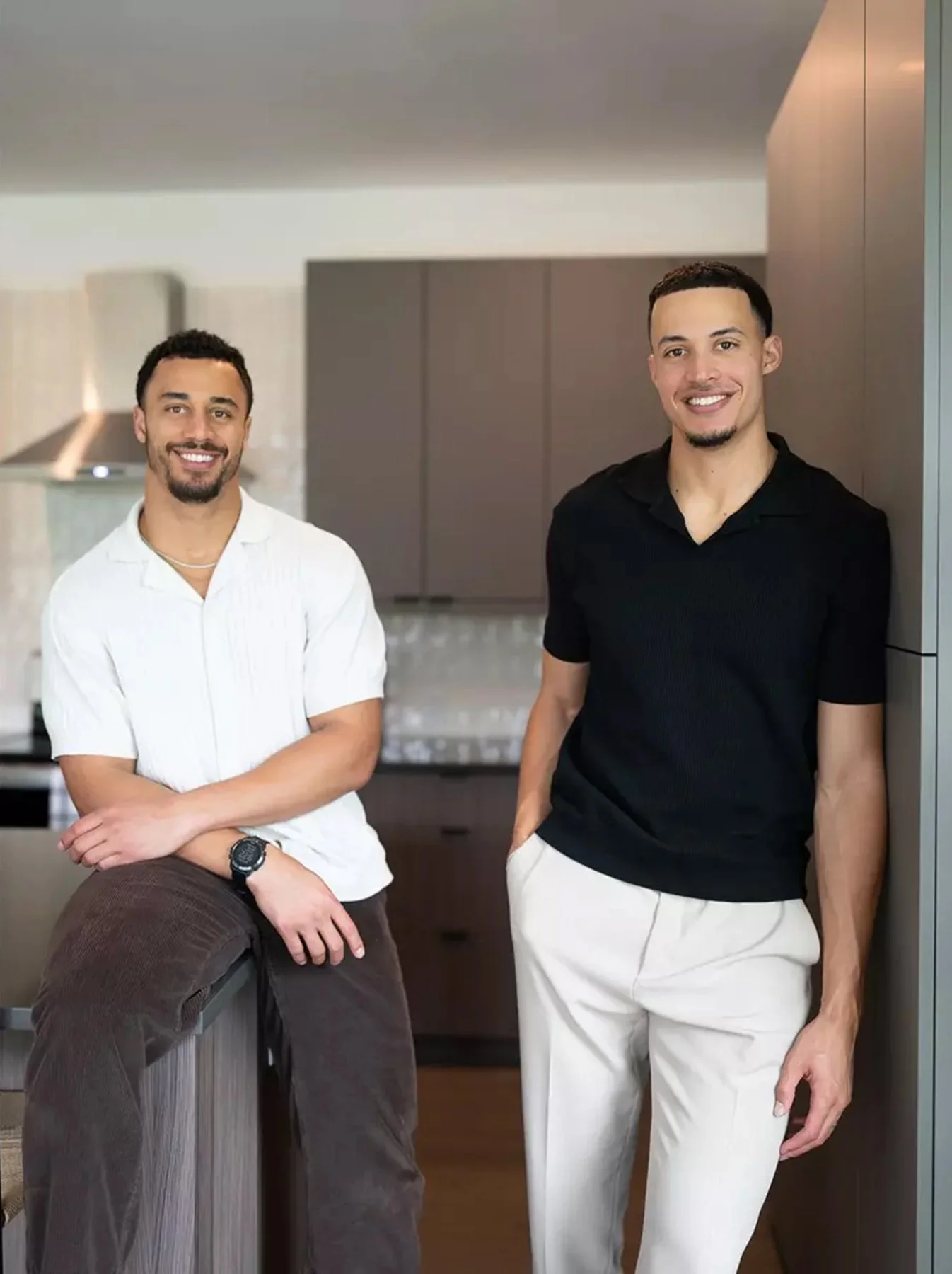 Two young men smiling in a modern kitchen, one wearing a white shirt and brown pants leaning on a counter, the other in a black shirt and white pants standing beside him.