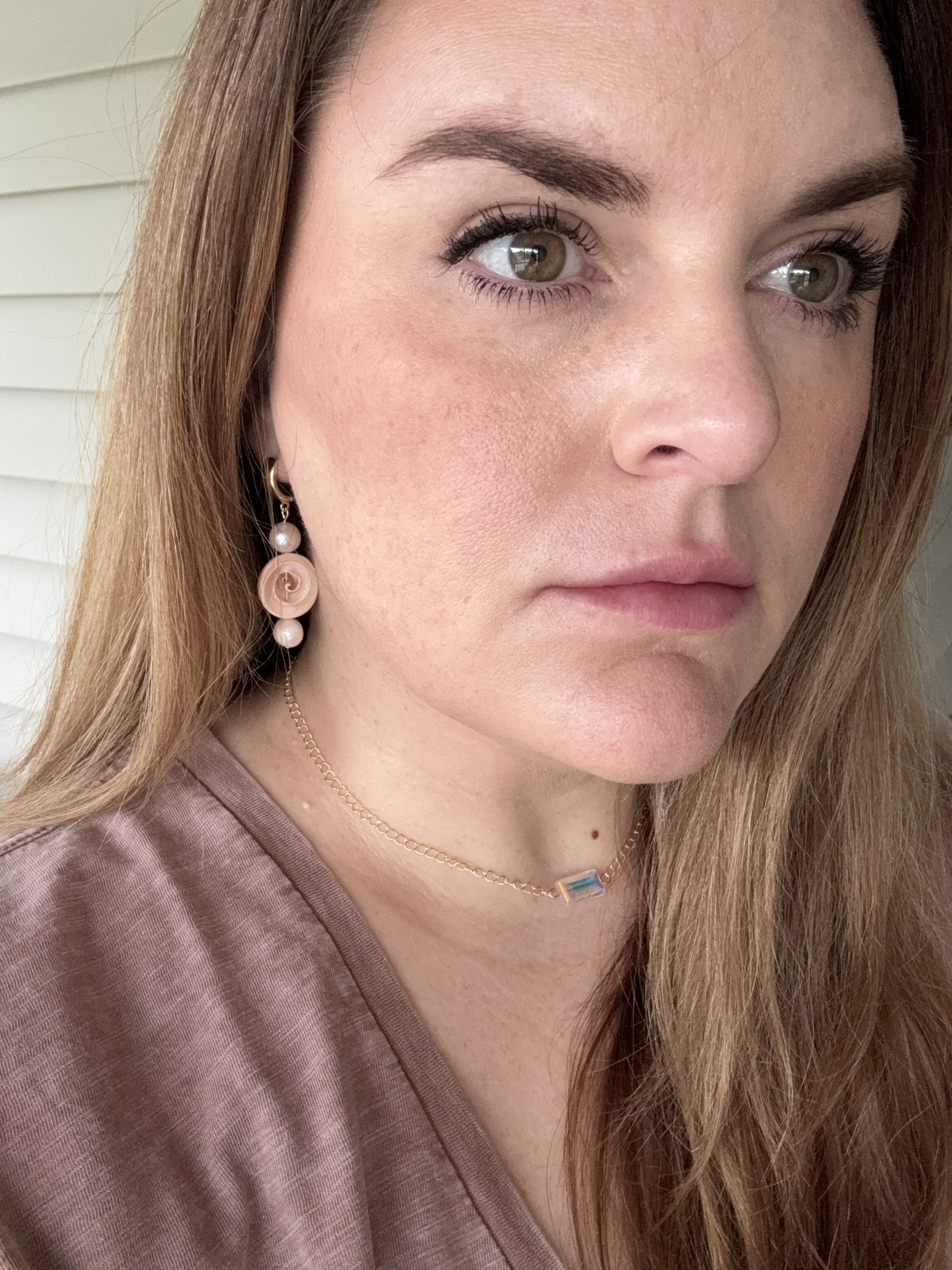 Close-up of a woman with light brown hair, light skin, and hazel eyes, wearing pink earrings and a delicate gold necklace with a rectangular pendant, dressed in a mauve top, standing in front of a white background.