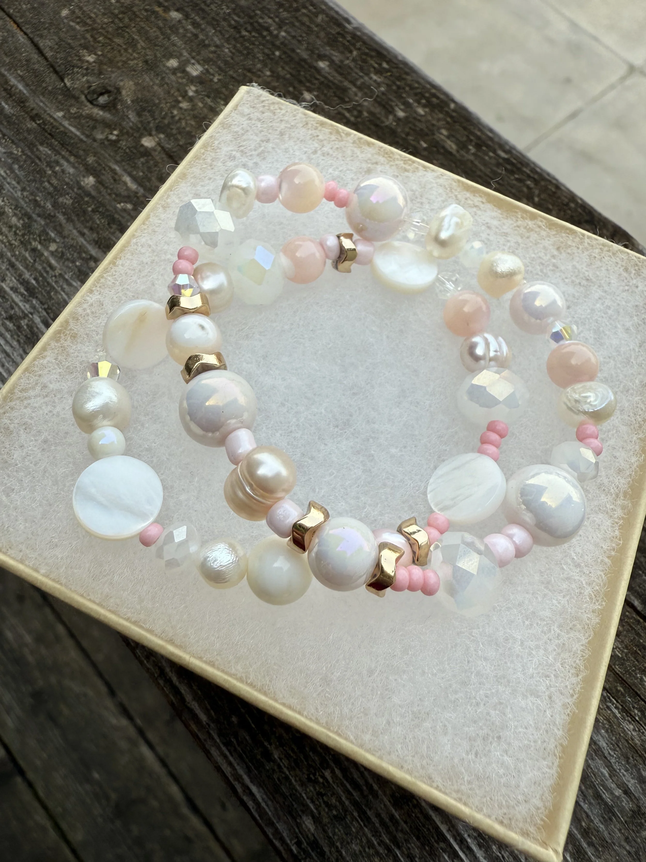Close-up of a jewelry box containing a pearl and bead bracelet on a textured light-colored surface, with a dark wooden background.