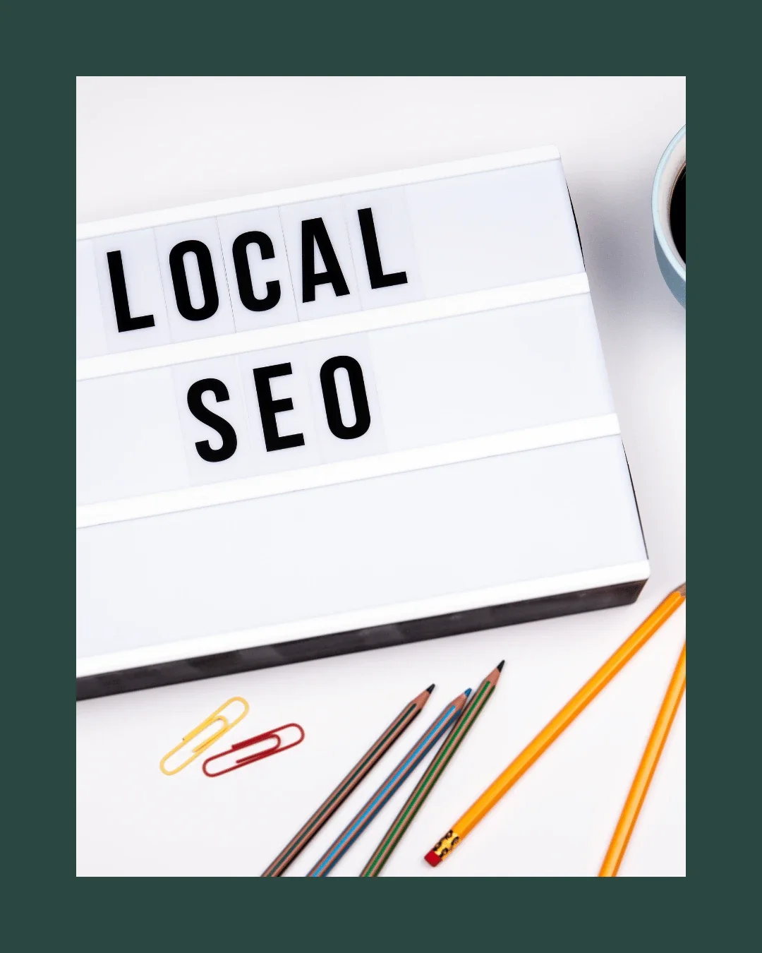 Lightbox displaying the words 'LOCAL SEO' surrounded by colored pencils, paper clips, and a cup of coffee on a white surface.