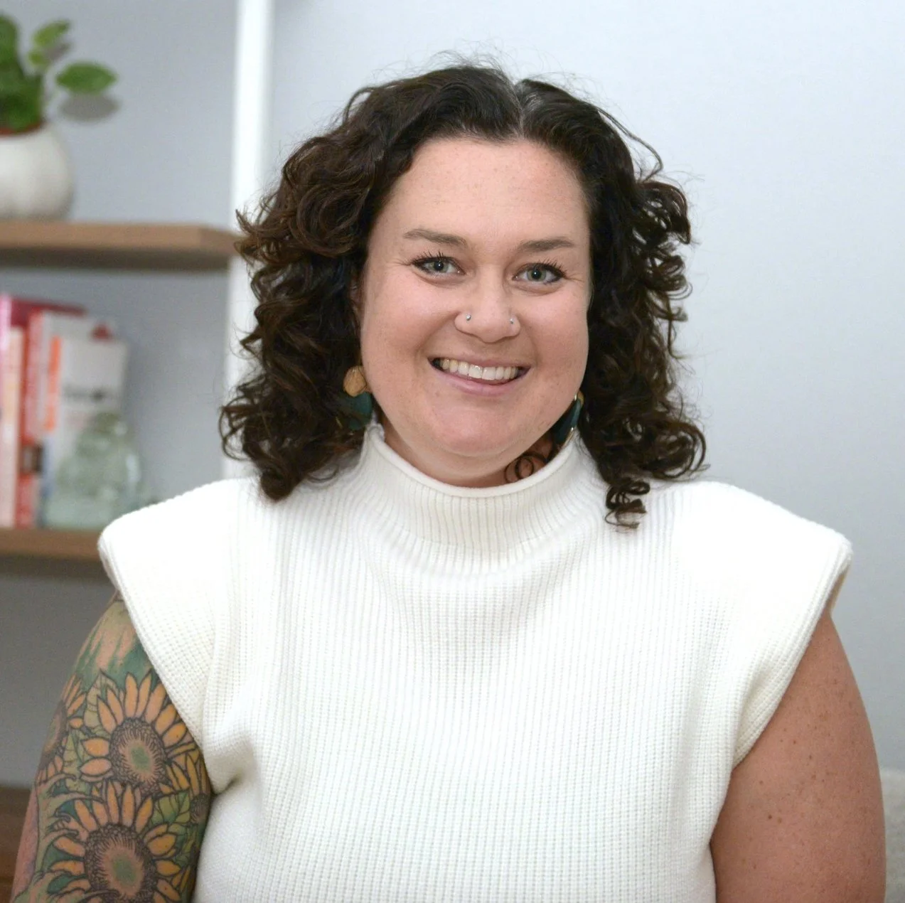 A smiling woman with curly dark hair, blue eyes, and tattoos on her left arm, wearing a white sleeveless turtleneck top, sitting indoors with a bookshelf and potted plant in the background.