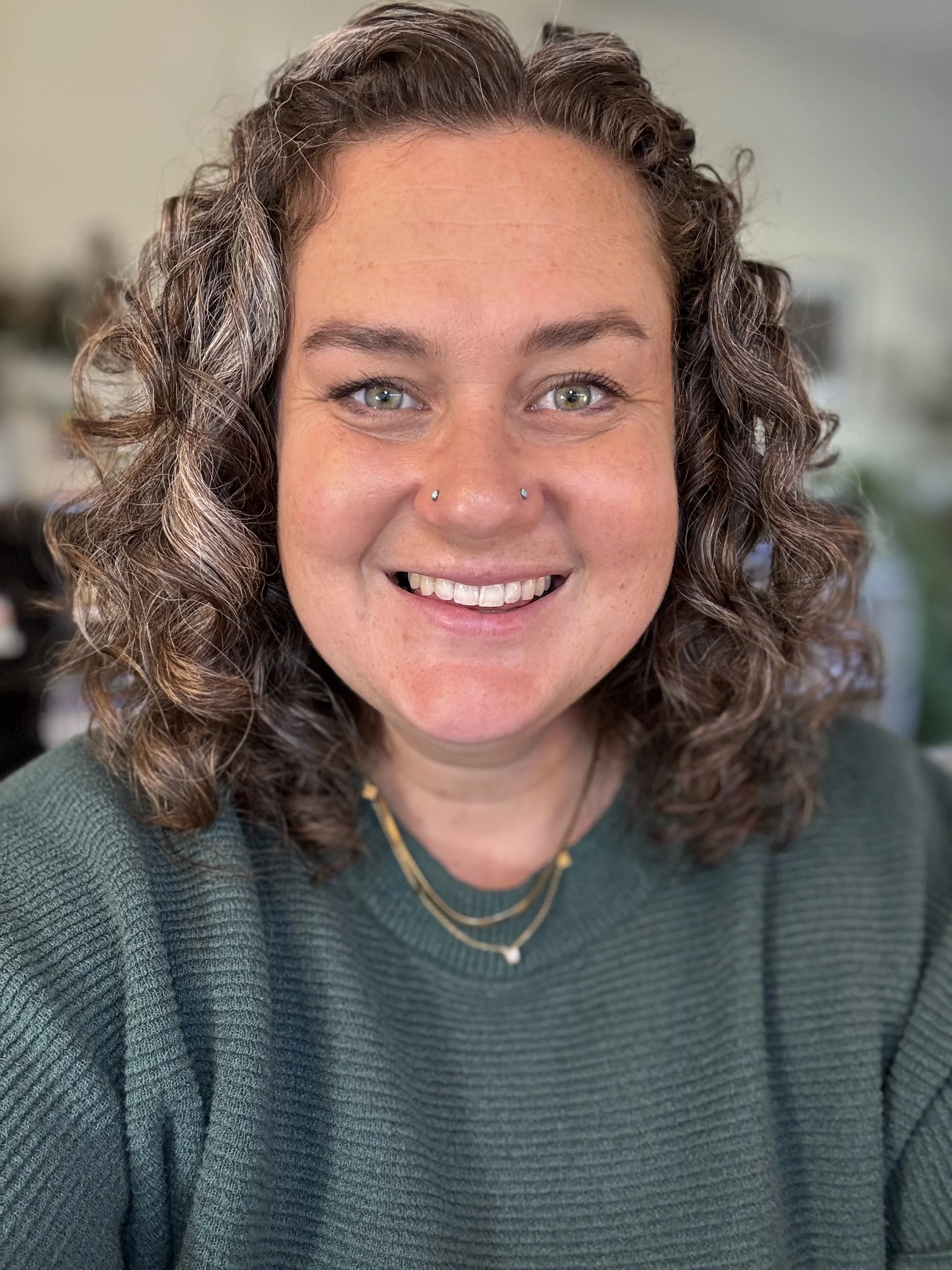 A smiling woman with curly brown hair, light blue eyes, and a nose piercing, wearing a dark green sweater and layered necklaces, in a well-lit room.