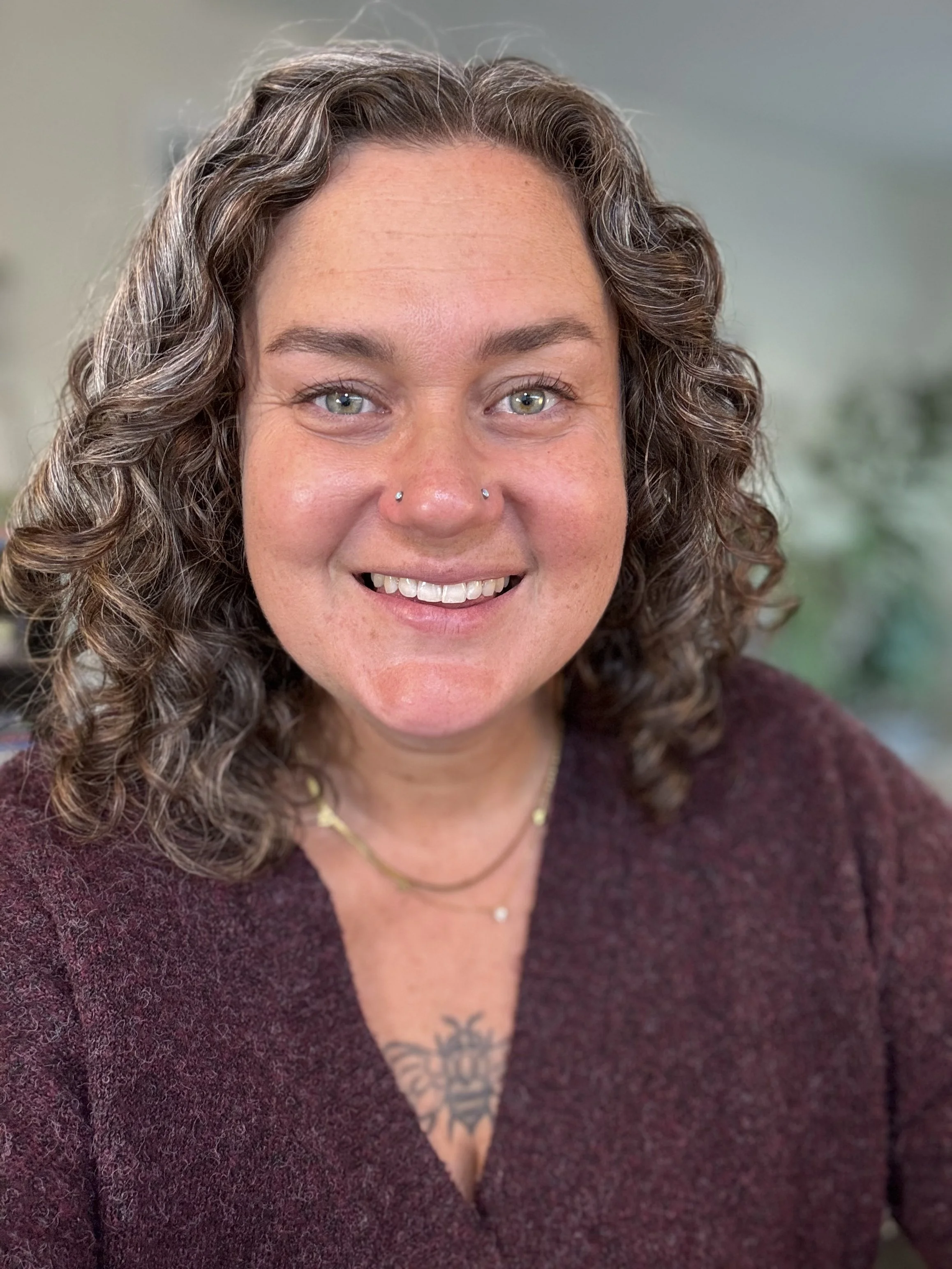 A woman with curly brown hair, blue eyes, and a nose piercing, smiling, wearing a maroon sweater and layered necklaces, with a tattoo of a bee visible on her chest.