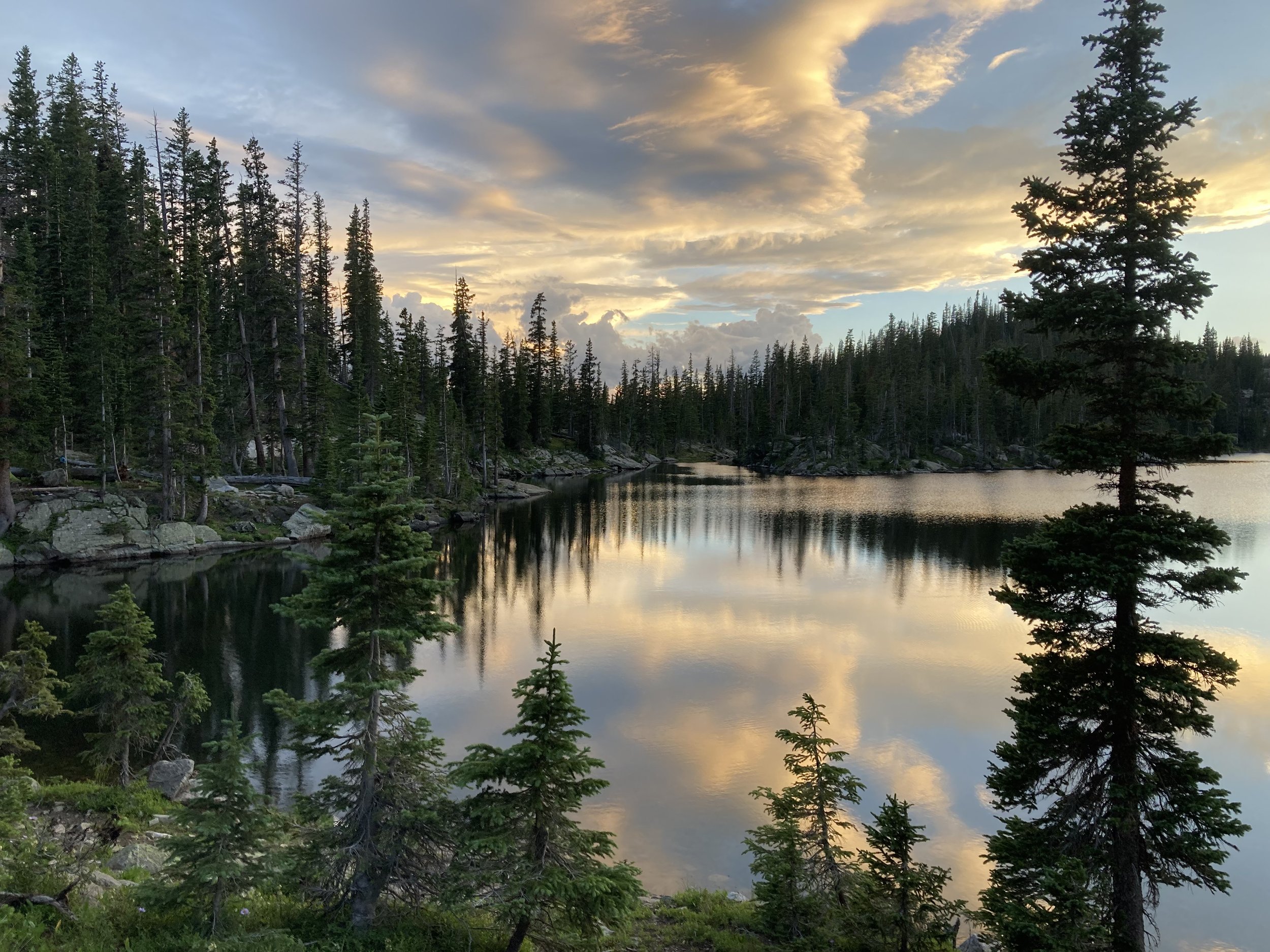 A serene lake surrounded by pine trees under a partly cloudy sky during sunset, with the water reflecting the sky's colors.