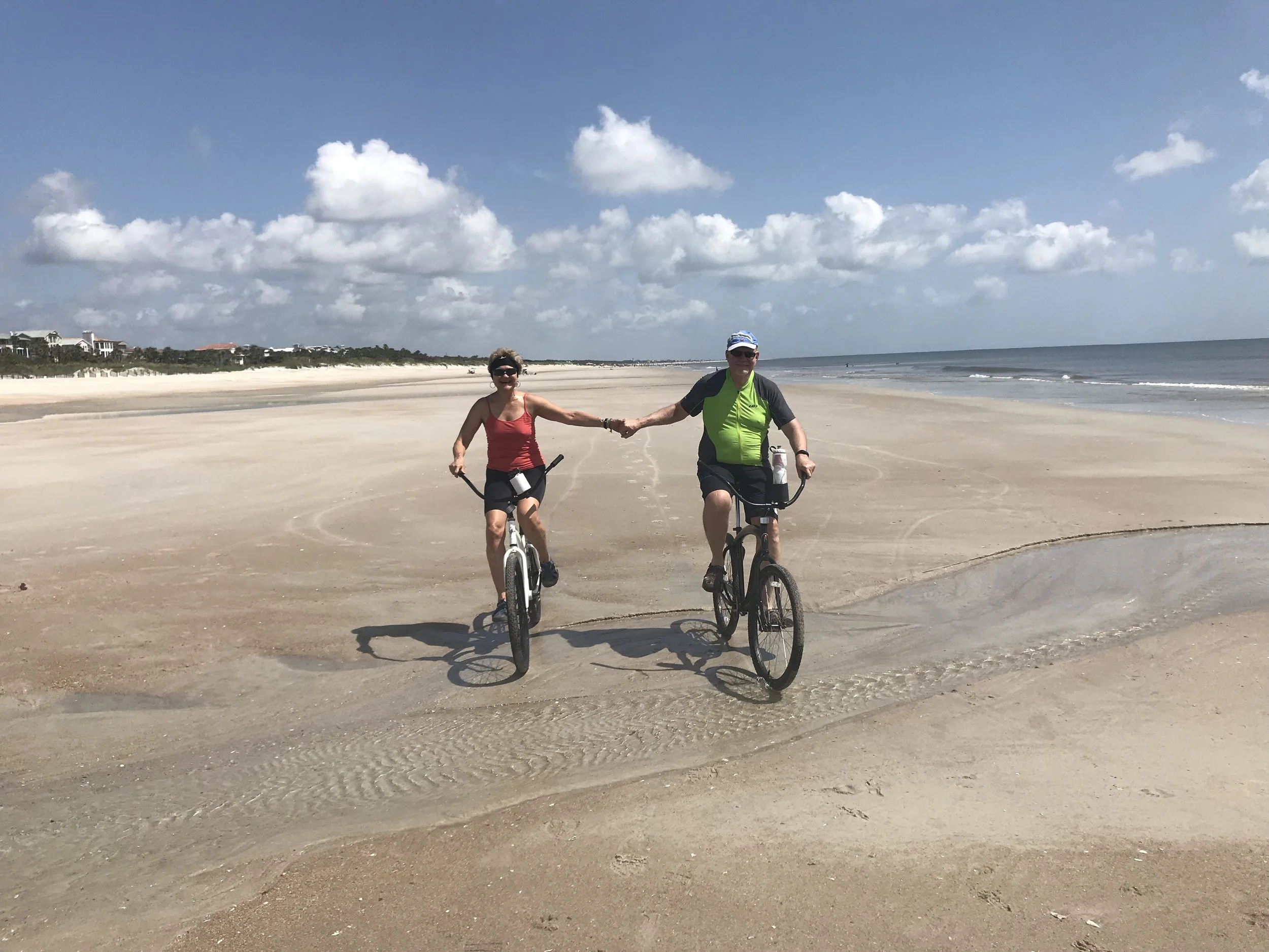 Two people riding bicycles on a sandy beach, holding hands, with white clouds and blue sky in the background.