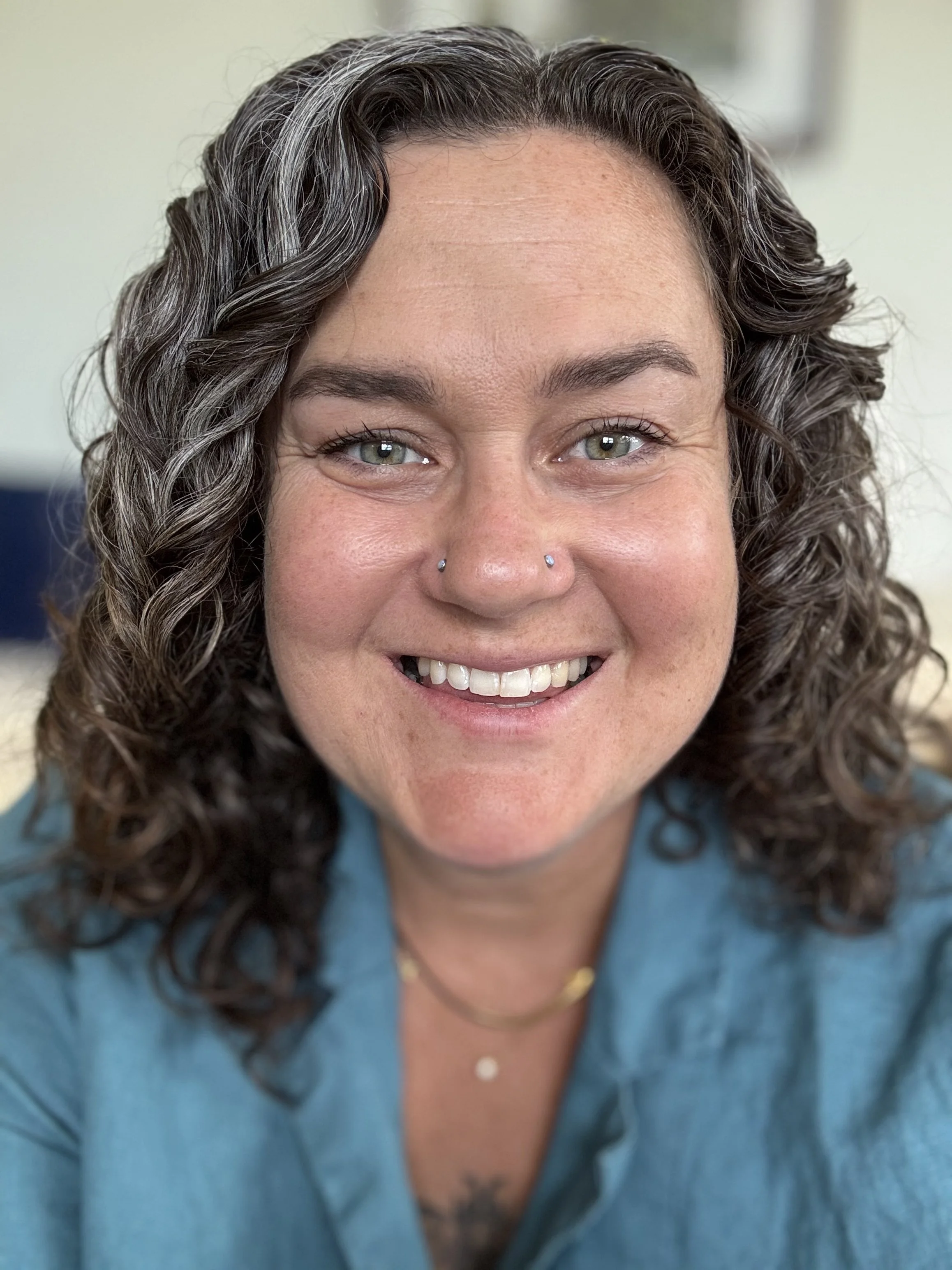 Close-up photo of a smiling woman with green eyes, curly dark hair, and nose piercings, wearing a blue shirt and gold jewelry.