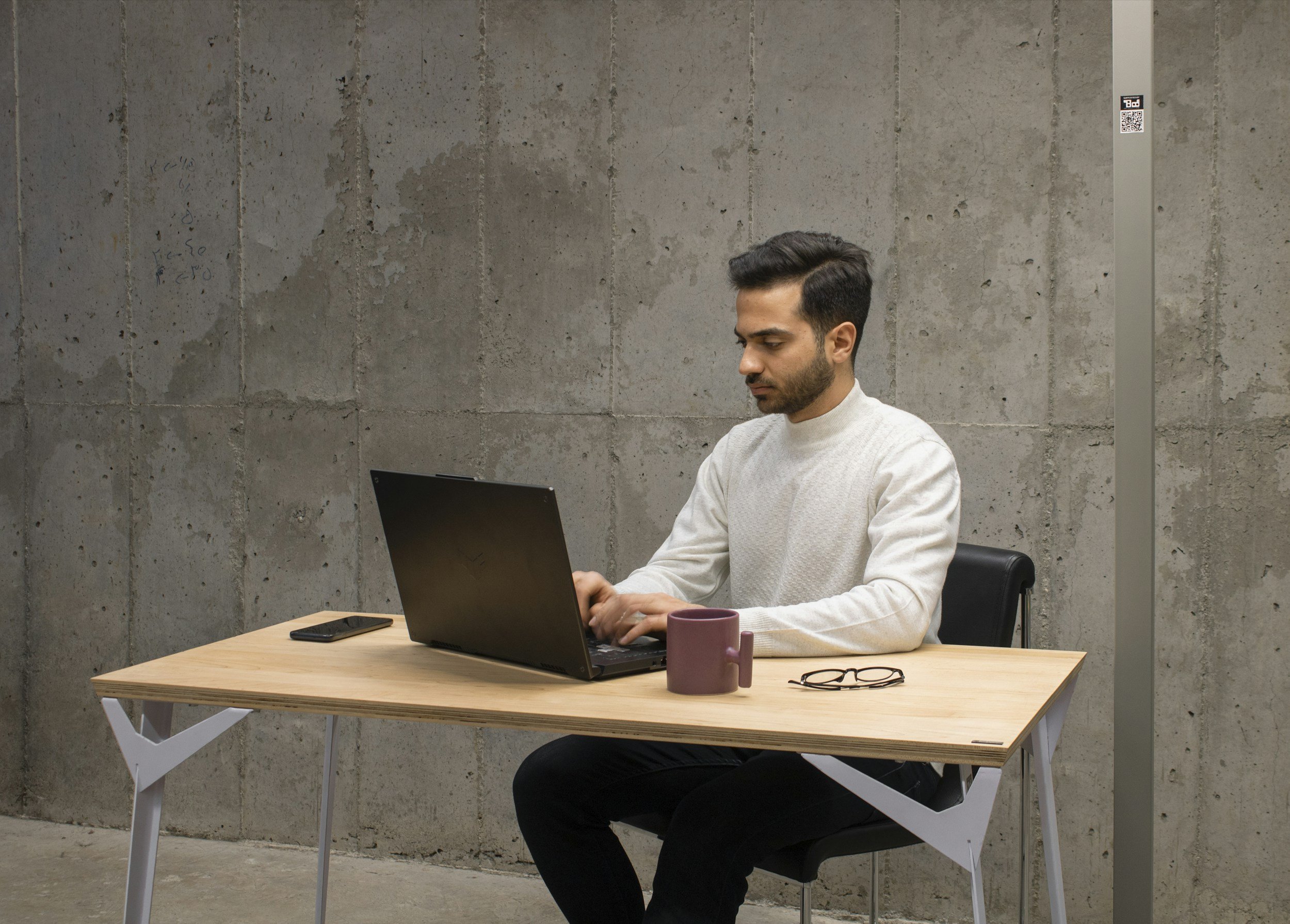 A man working on a laptop at a wooden desk in an industrial-style room with concrete walls. A smartphone and glasses are on the desk, and there are two mugs in front of him.