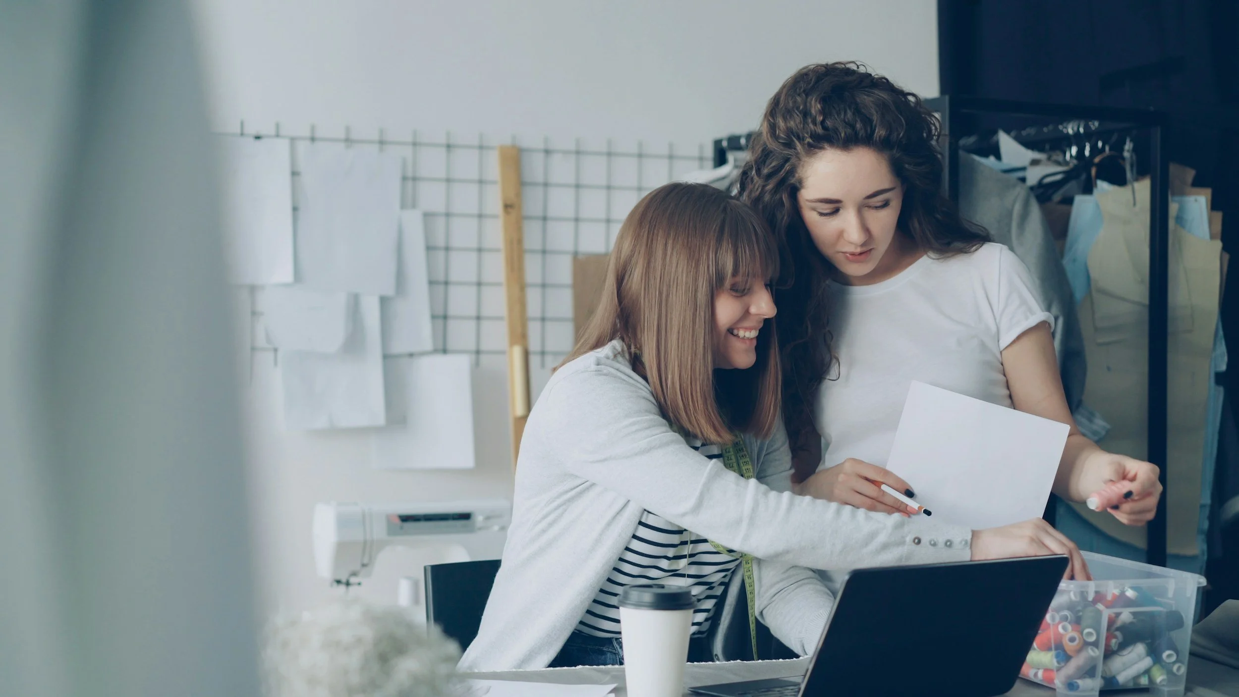 Two women working together at a sewing or fabric workspace, looking at a piece of paper with fabric patterns or design instructions, surrounded by sewing supplies and fabric in a bright, organized studio.