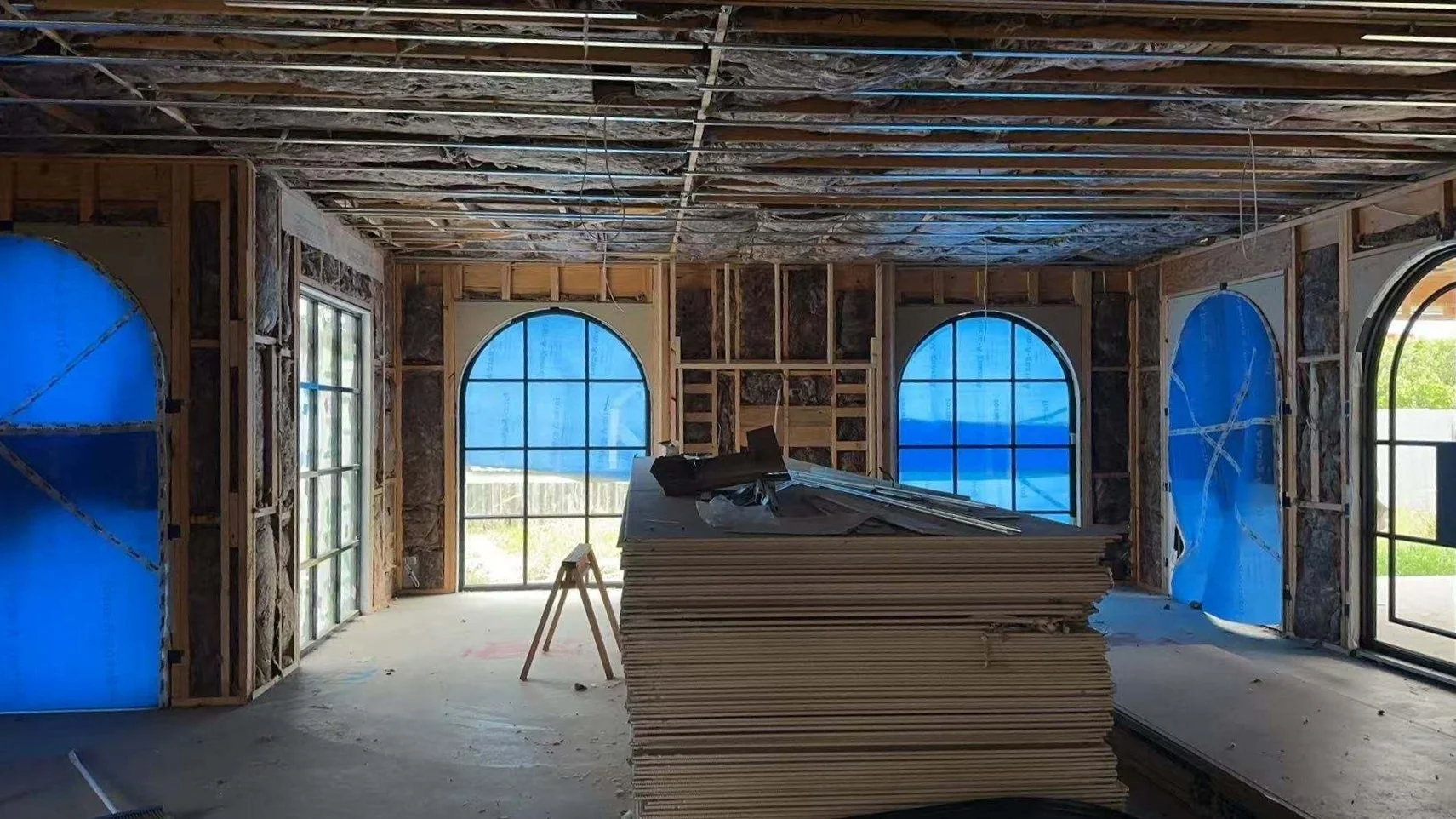 Interior of a house under construction with framed walls, blue protective coverings over the windows and doors, and construction materials stacked in the center.