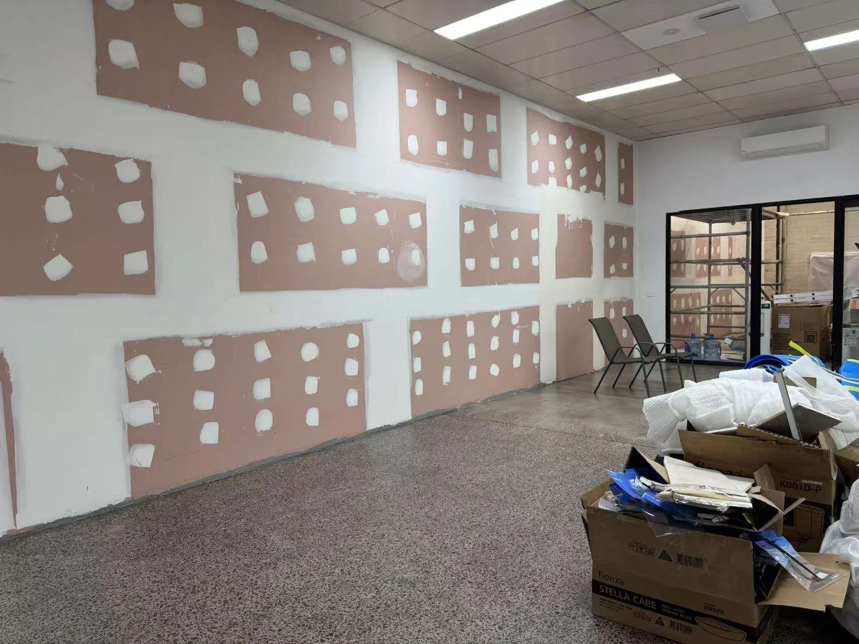 An indoor space under renovation with patched drywall on the walls, a tiled ceiling, and a windowed door. Construction materials and tools are in a box, and two chairs are against the wall.