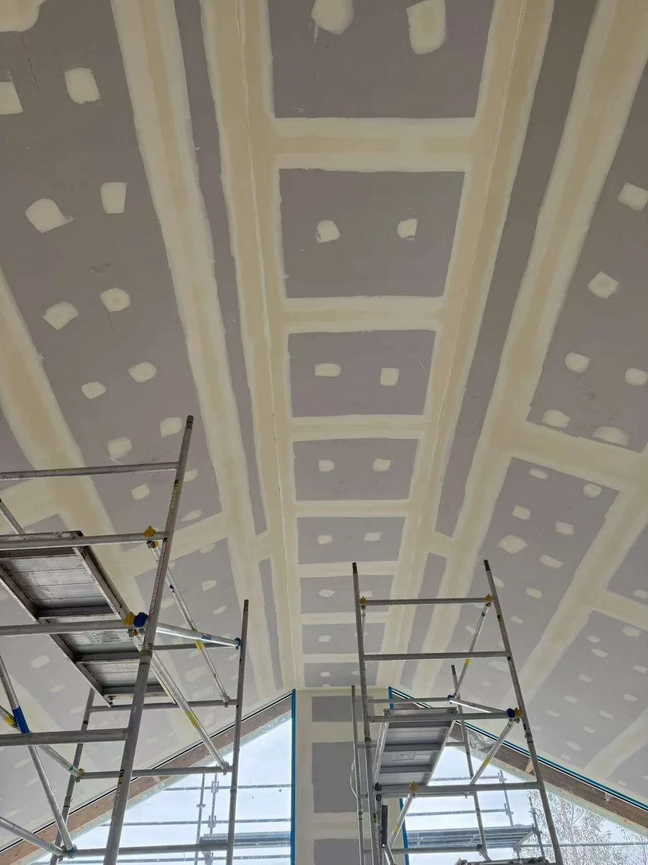 Ceiling with drywall and joint compound being applied, with scaffolding and ladders in a construction site.
