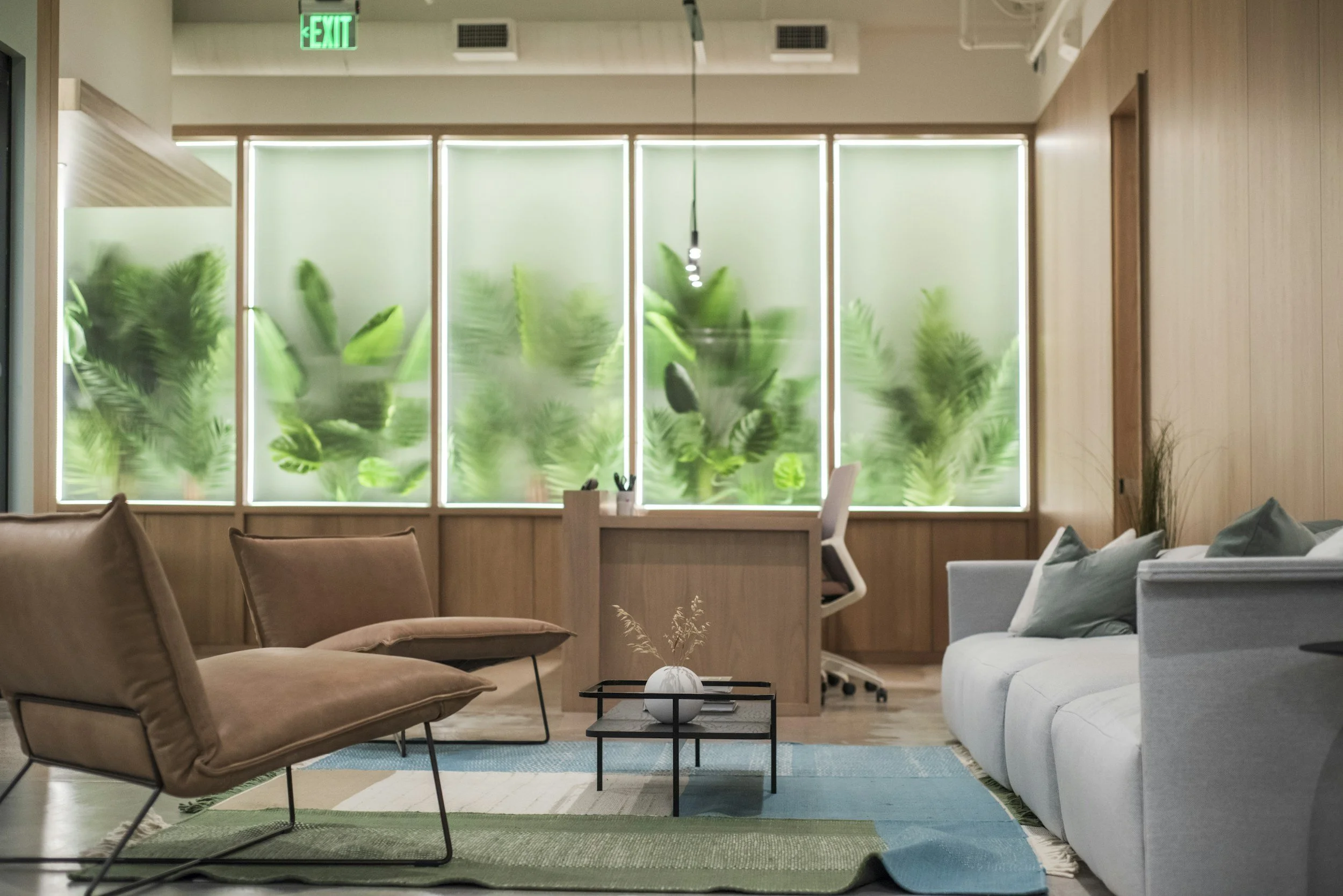 Modern office waiting area with beige and white couches, a small black coffee table with a white vase and dried plants, a wooden front desk, and a frosted window with green plants behind it. Green exit sign is visible on the ceiling.
