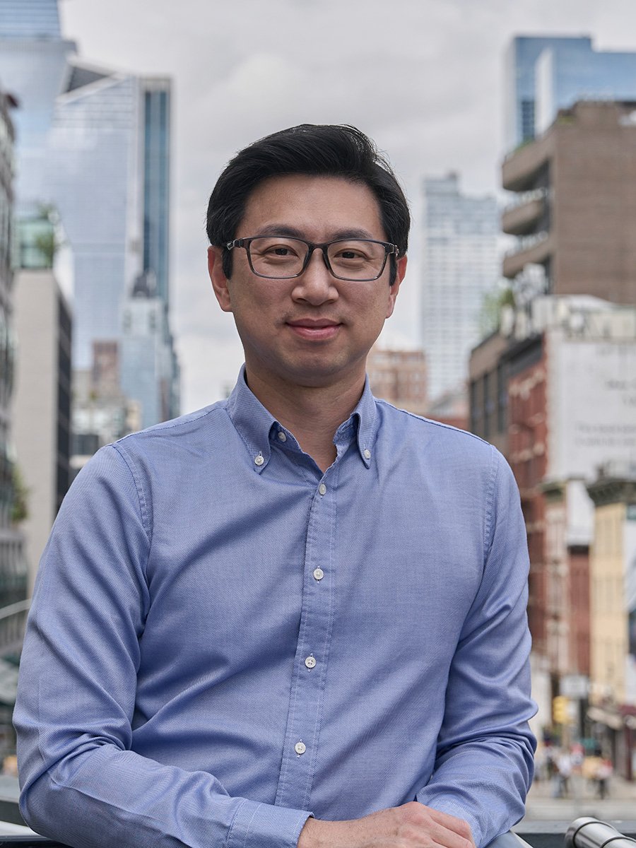 A man with black hair, glasses, wearing a blue button-up shirt, standing outdoors in an urban city with tall buildings and cloudy sky in the background.